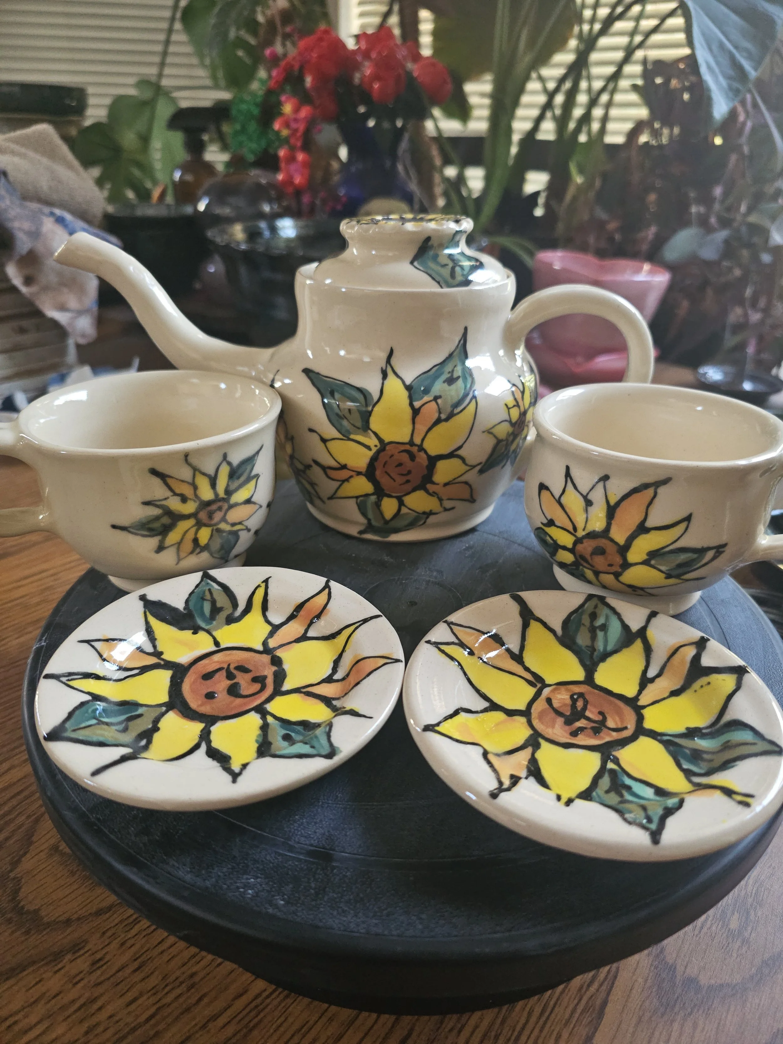 A decorative ceramic teapot and three matching cups with hand-painted yellow sunflowers and green leaves, placed on a black tray.