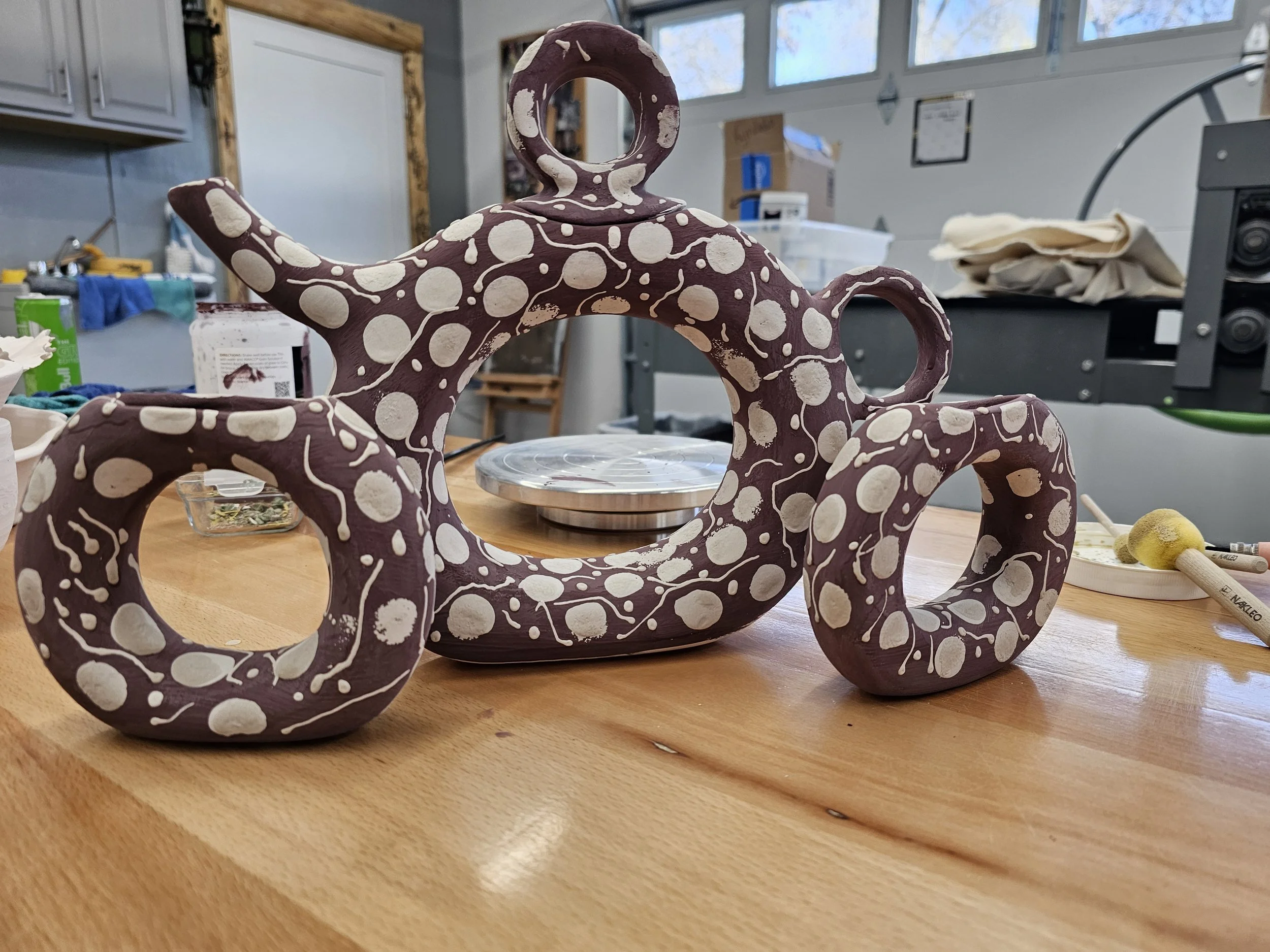 A decorative ceramic wheel-thrown teapot set with a base color and white polka dots and details, placed on a wooden table in an indoor workshop setting.