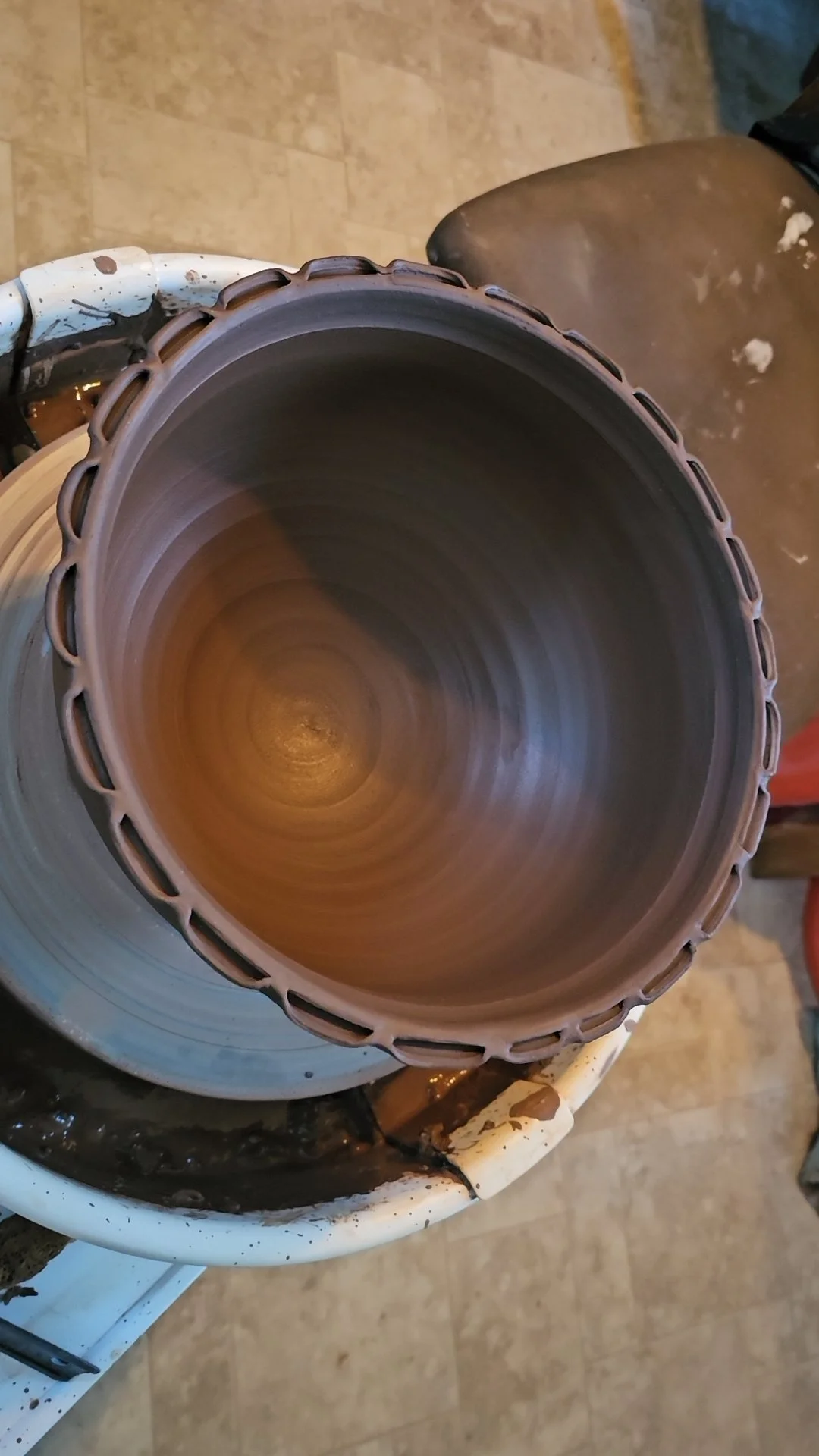 Inside view of a brown ceramic pottery piece being shaped on a pottery wheel, with the potter's workspace visible around.