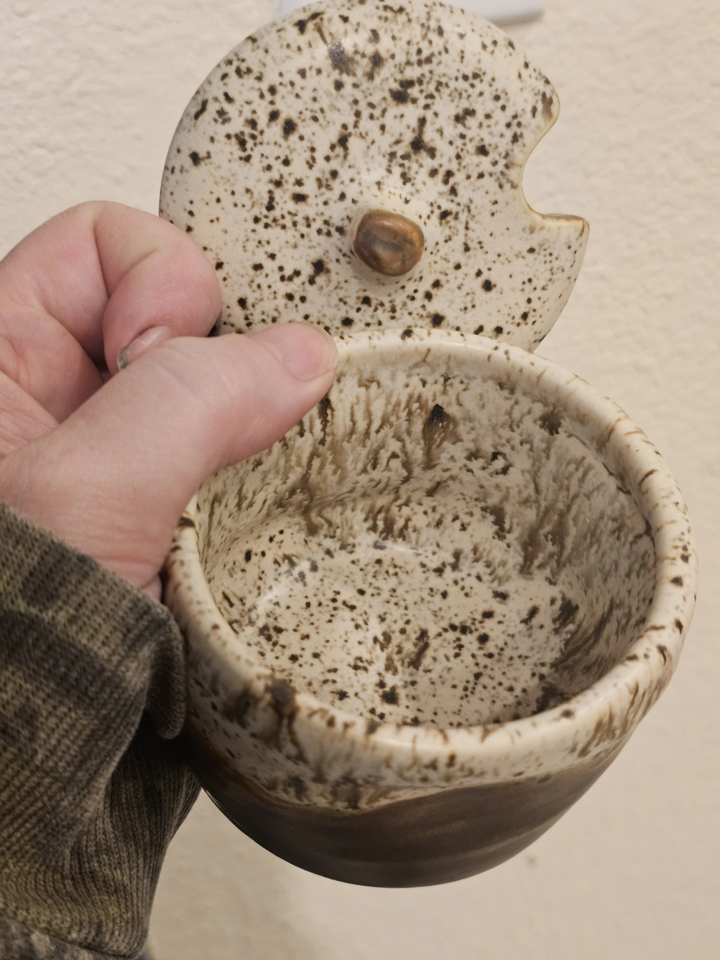 Close-up of a small ceramic container with a lid, both decorated with a speckled pattern, held by a person's hand.