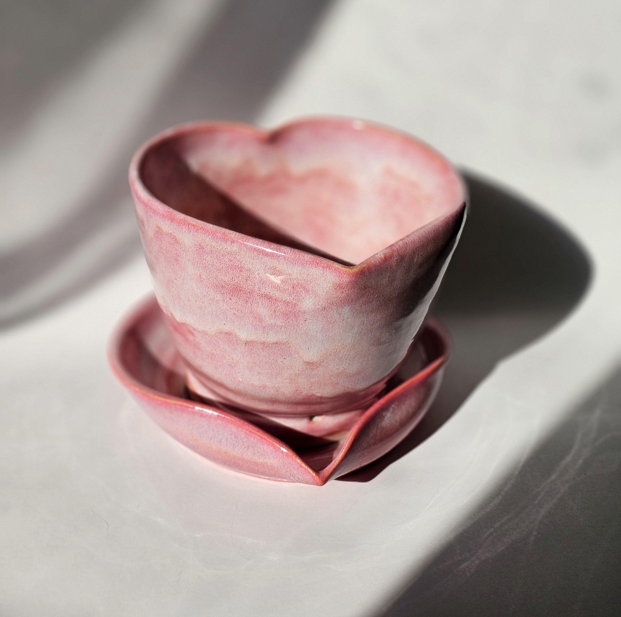 Close-up of a pink heart-shaped object made of a ceramic  material, with a hollow interior, placed on a white surface with shadows cast nearby.