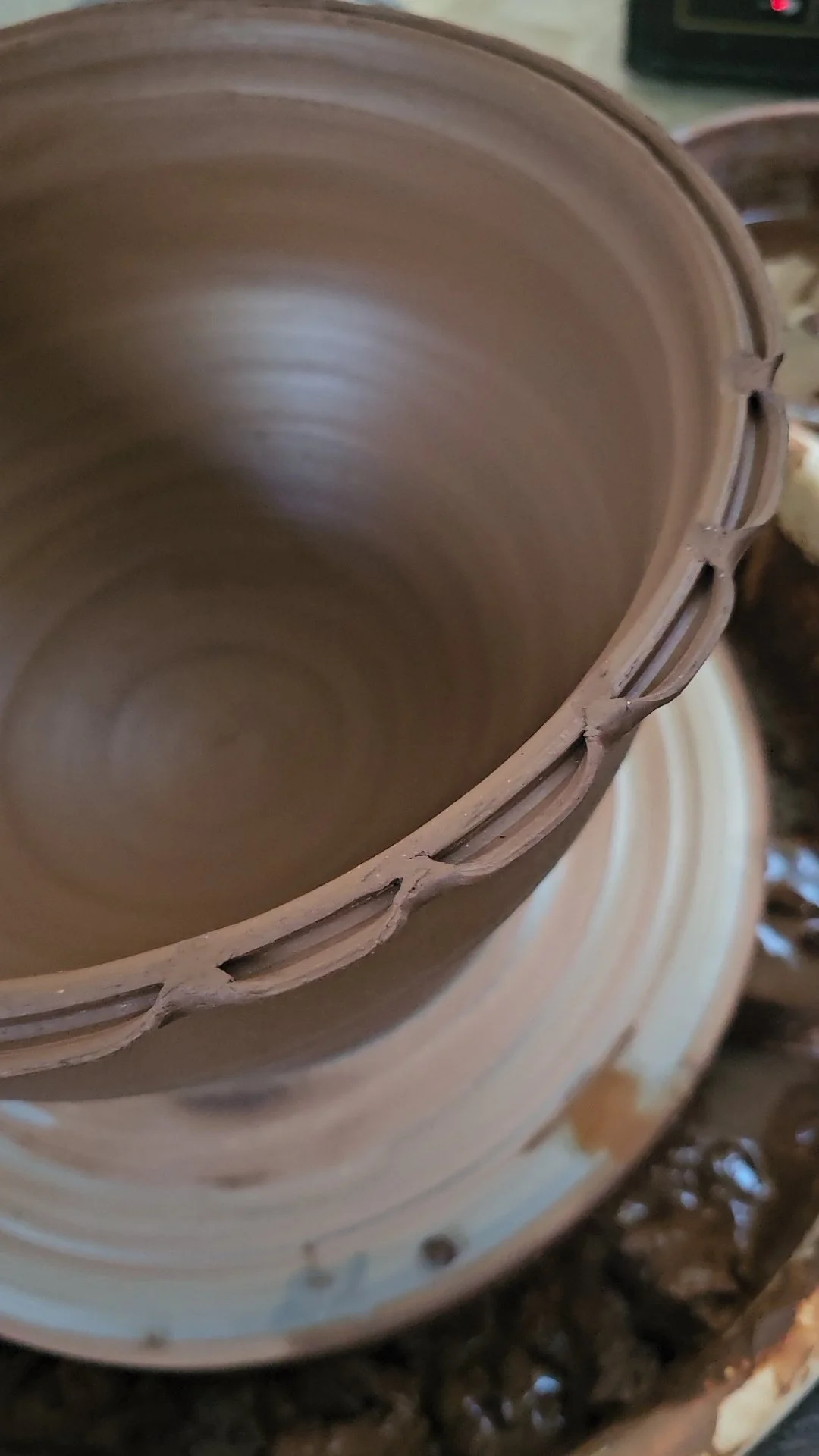 Close-up of the inside of a brown clay or ceramic pot with a textured rim, placed on a dark surface with some blurred objects nearby.