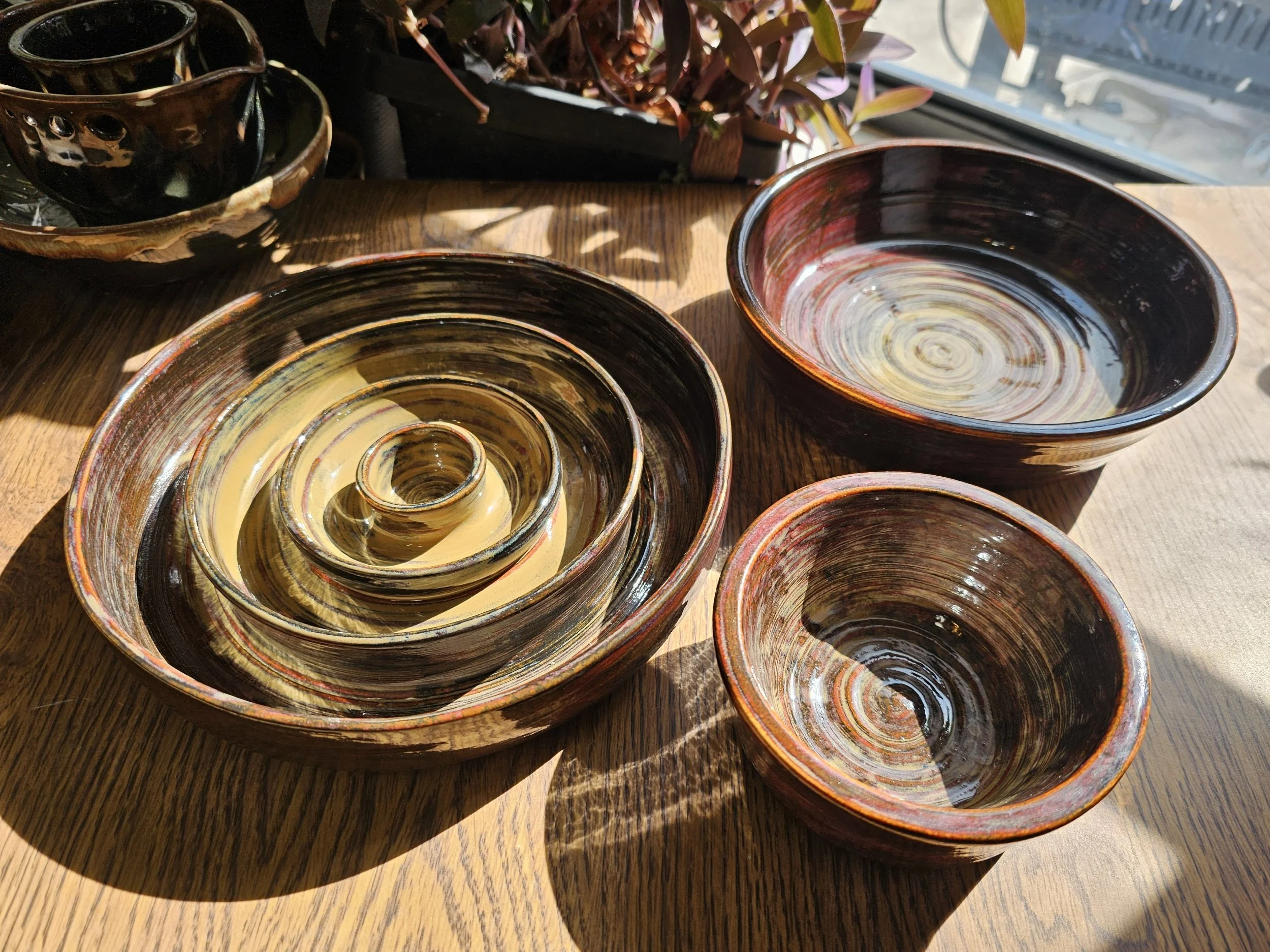 Colorful wheel-thrown ceramic bowls of various sizes stacked and placed on a wooden table near a window. Kyndal's Clay Creations