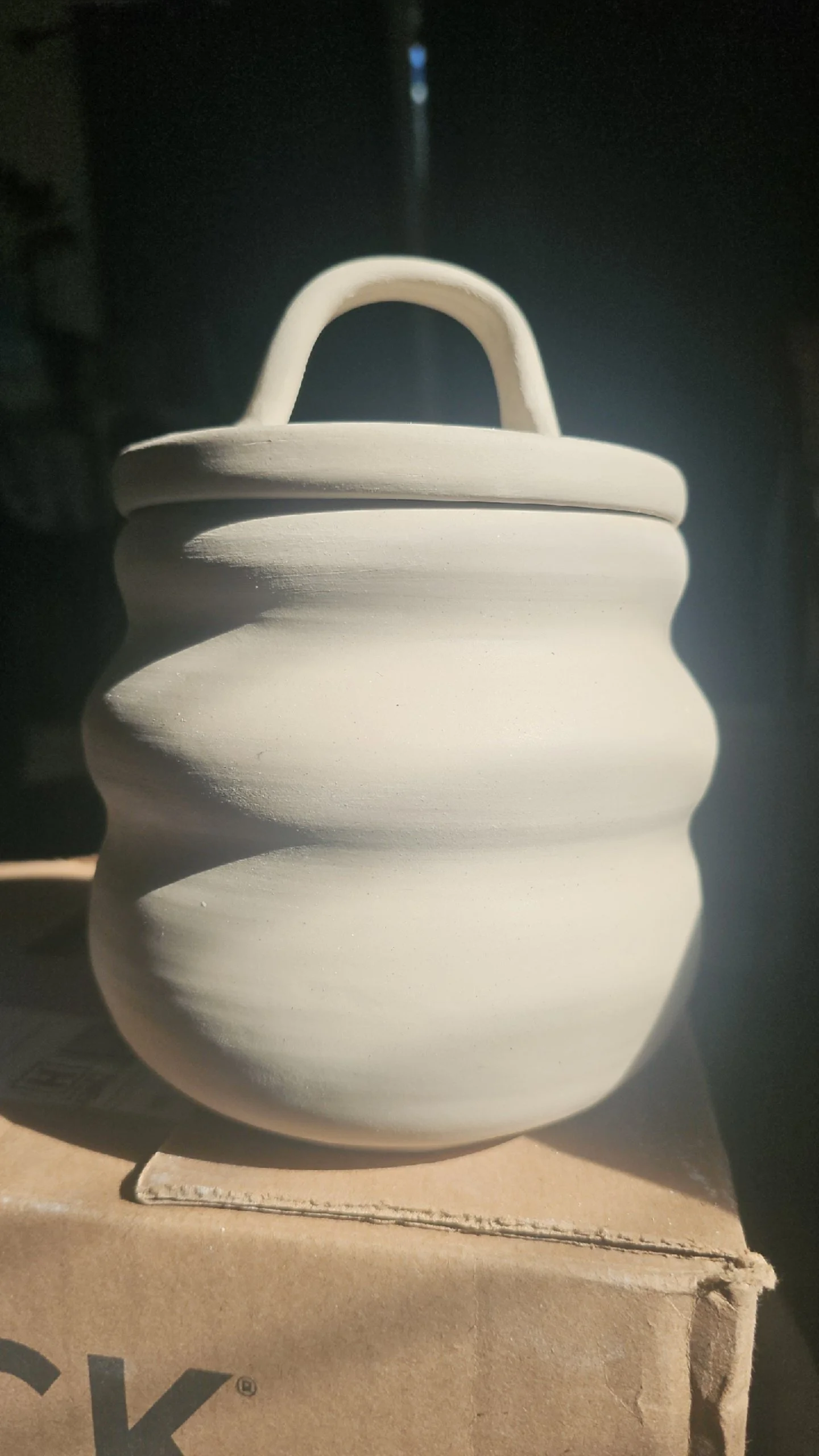 Unfinished white ceramic pot with a handle, sitting on a cardboard box, with a dark background and a light source creating shadows.