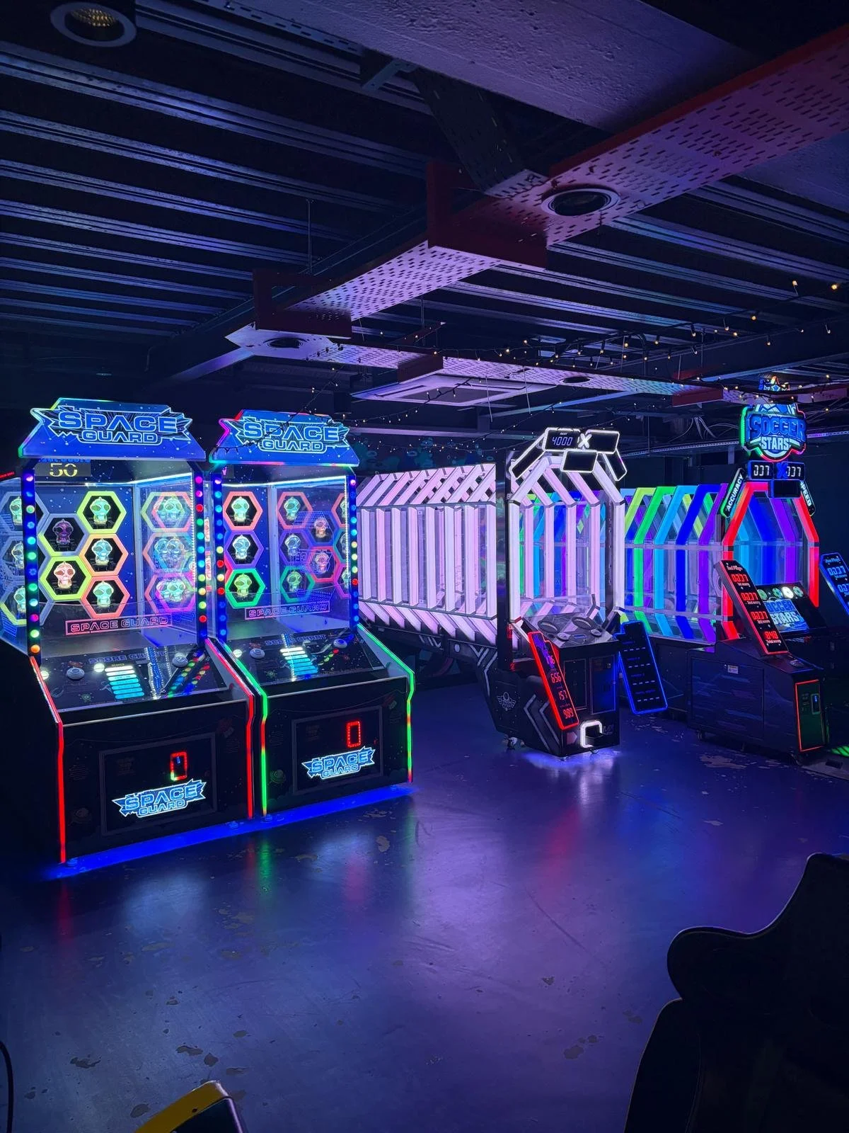 Colorful arcade games with neon lights, including space-themed and soccer-themed machines, in a dark gaming room.