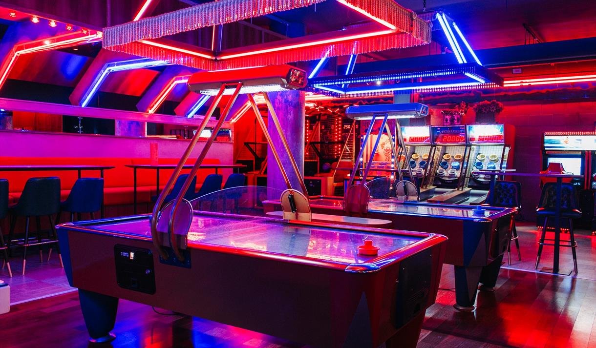 Red, blue, and purple neon-lit arcade with air hockey tables, pinball machines, and seating area.