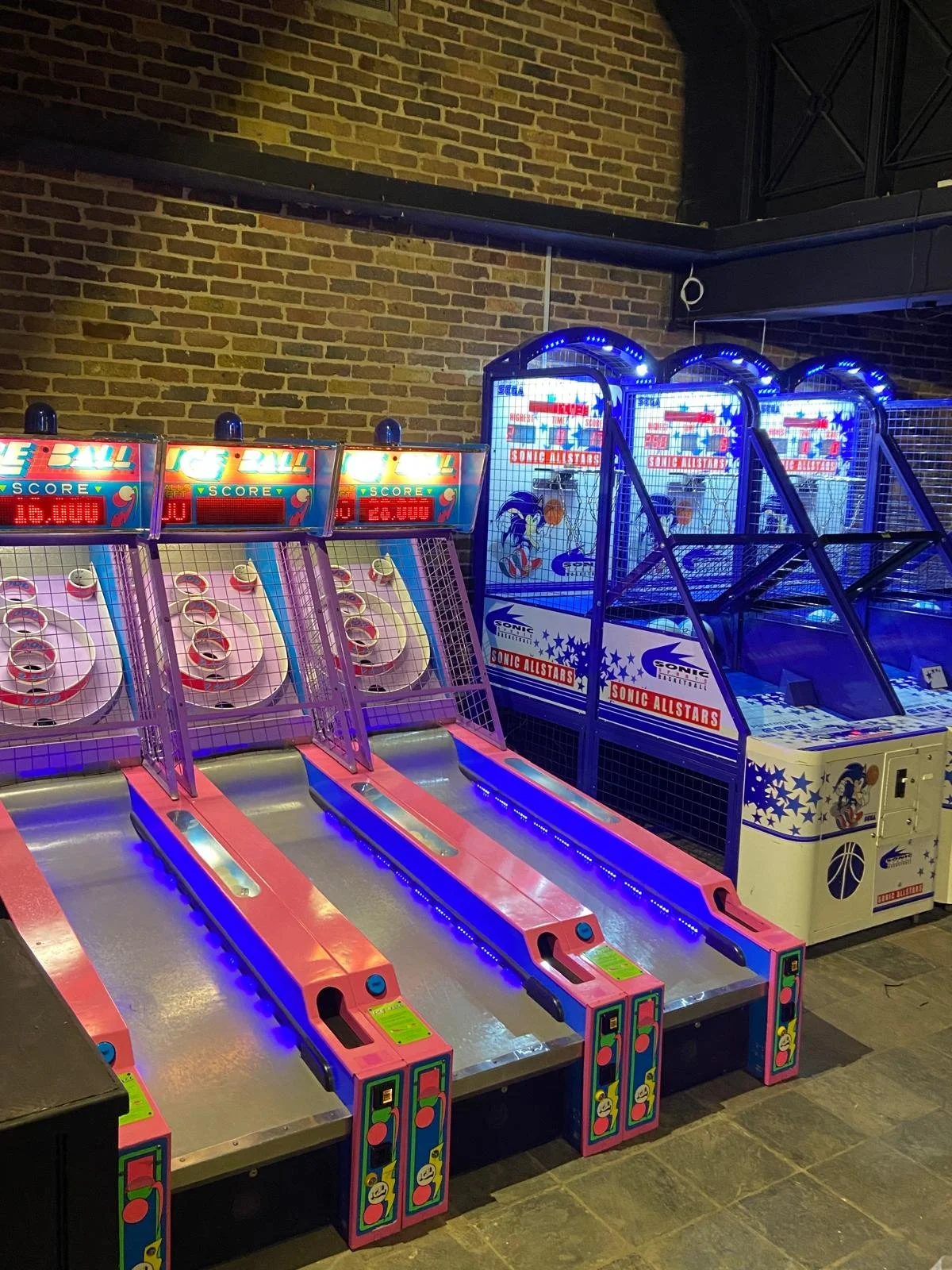 Three skee-ball arcade machines and two basketball arcade games in a game room with a brick wall background.