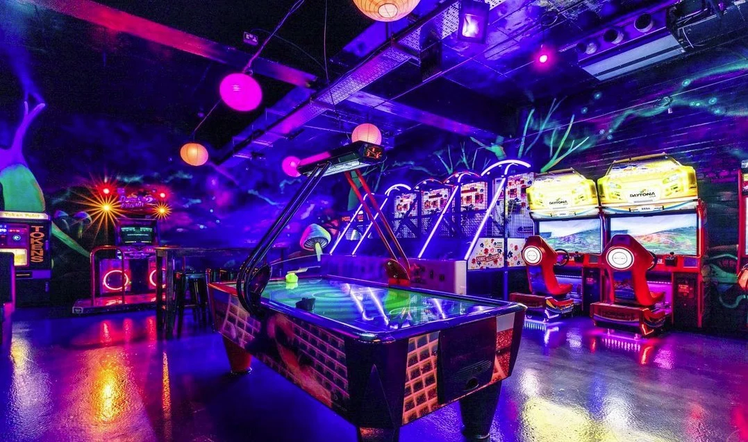 An arcade with neon lights featuring an air hockey table, racing game chairs, and various arcade machines.