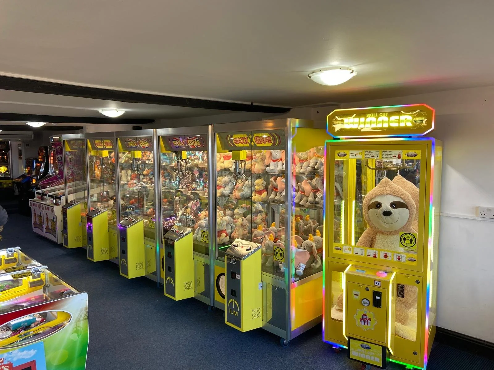 A row of arcade claw machines filled with plush toys, with a yellow claw machine on the right featuring a sloth plush toy wearing a bear costume.