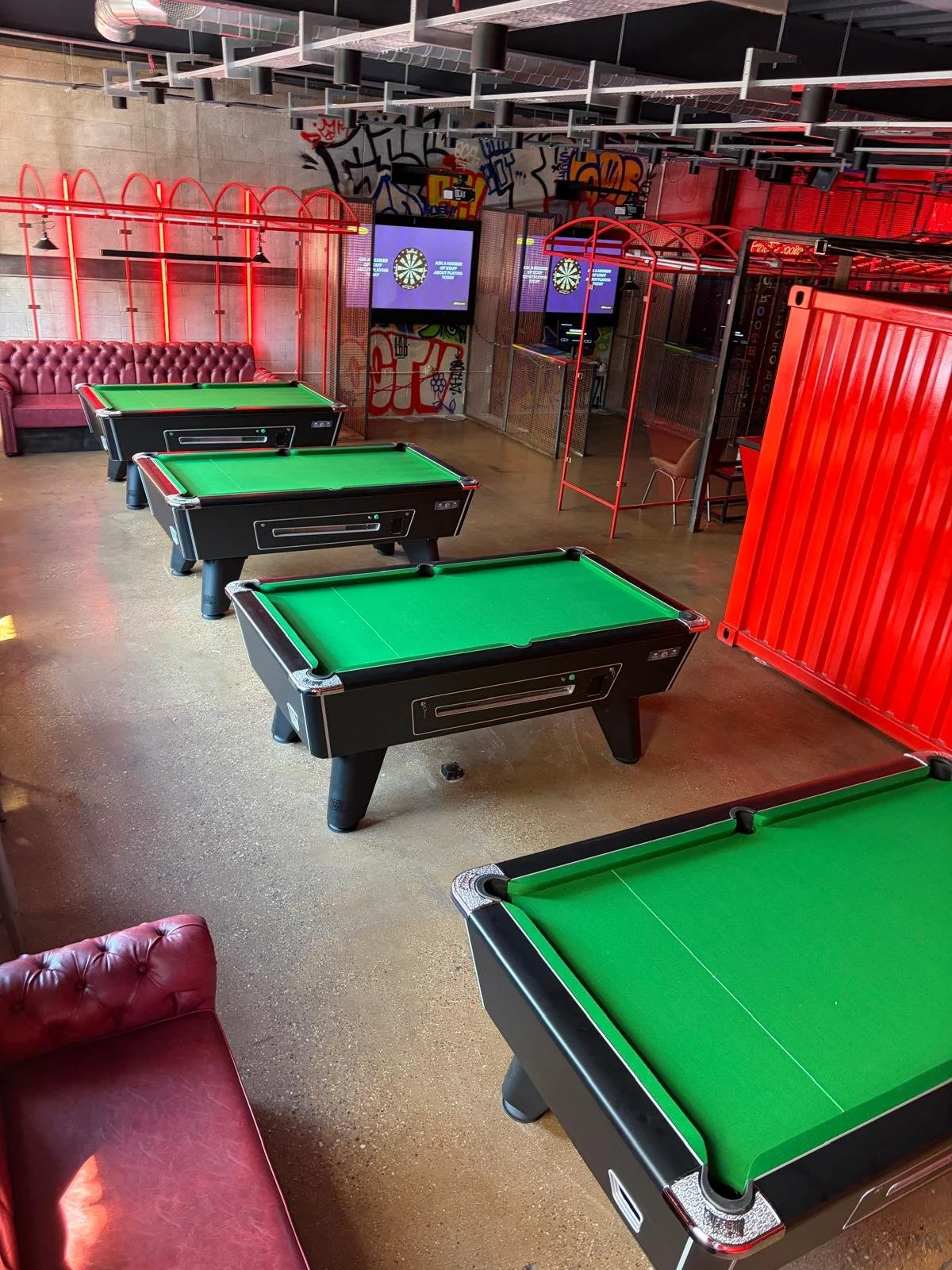 Indoor game room with three black pool tables with green felt, red lighting and graffiti art on the walls, a red sofa, and two digital dartboards.
