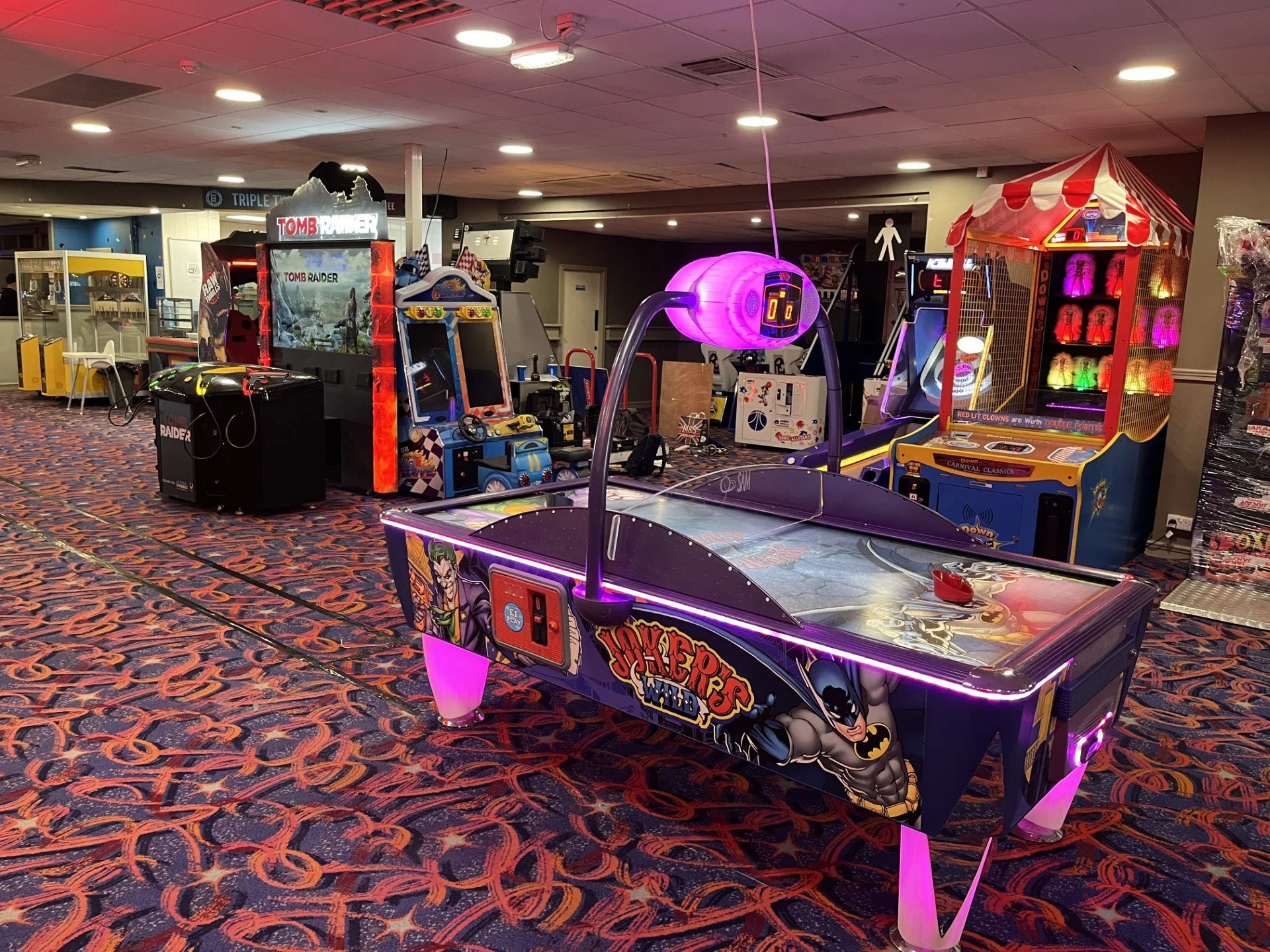 Inside an arcade featuring various video games and amusement machines, including an air hockey table with Joker and Batman graphics, a purple-lit tunnel, a claw machine with colorful prizes, and other game cabinets with vibrant neon lighting. The floor has a patterned carpet and ceiling lights illuminate the area.