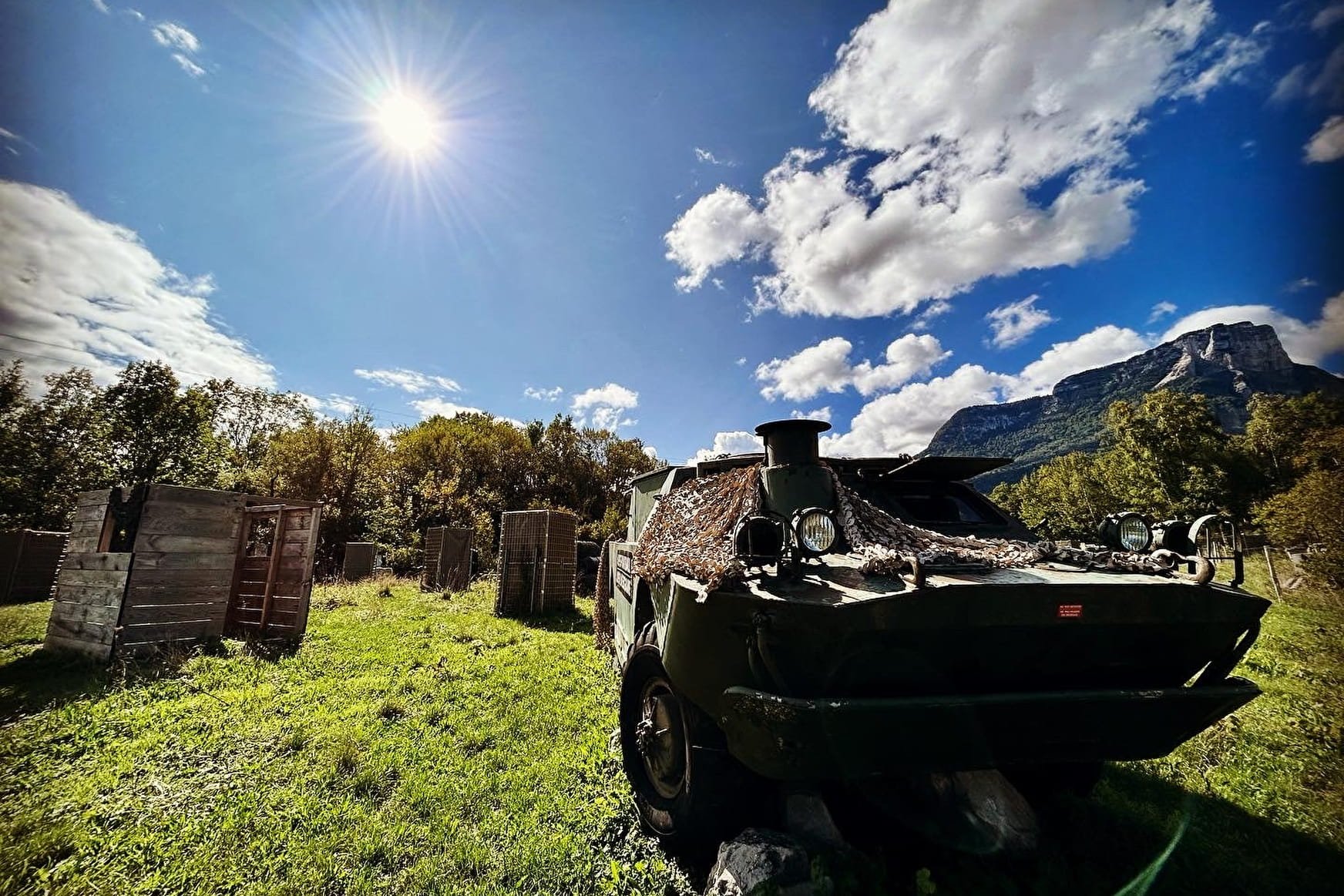 Une voiture ancienne et rouillée est stationnée dans un champ vert en plein jour, sous un ciel bleu avec des nuages blancs, avec des montagnes en arrière-plan et des arbres autour.
