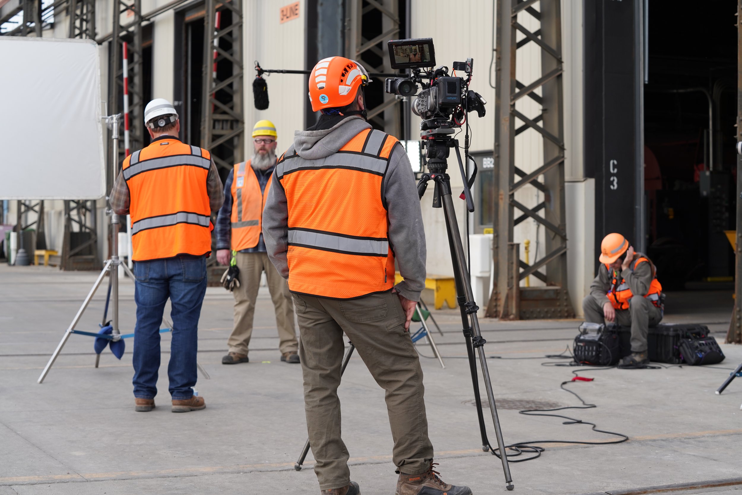 Crew members wearing safety vests and helmets setting up camera equipment outdoors near a building with scaffolding.