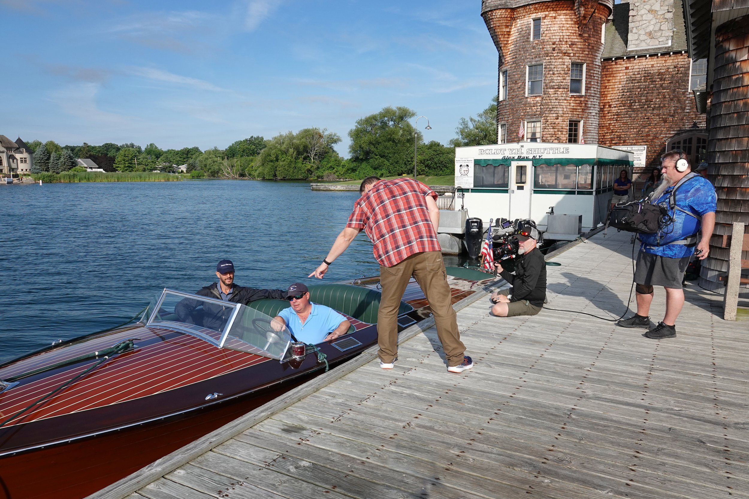 People on a dock preparing to board a vintage wooden speedboat near a river, with a historic building and trees in the background.