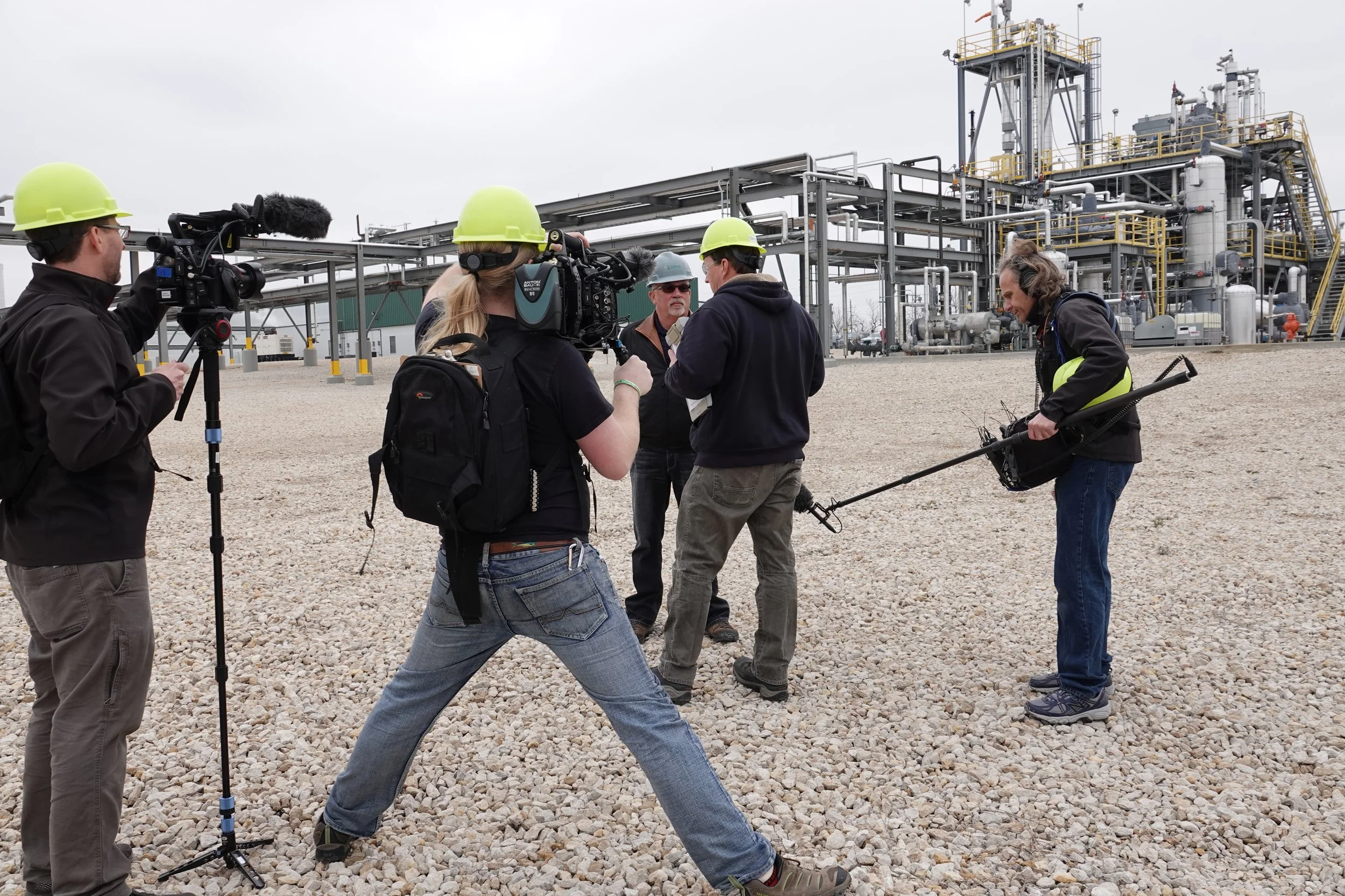A film crew is filming a documentary at an industrial plant, with multiple crew members wearing yellow safety helmets, filming equipment, and one person holding a microphone boom, while the plant has complex pipes and structures in the background.