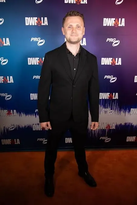 A man in a black suit standing on a red carpet in front of a backdrop with logos for DWE LA, PRO, and other sponsors at an event.