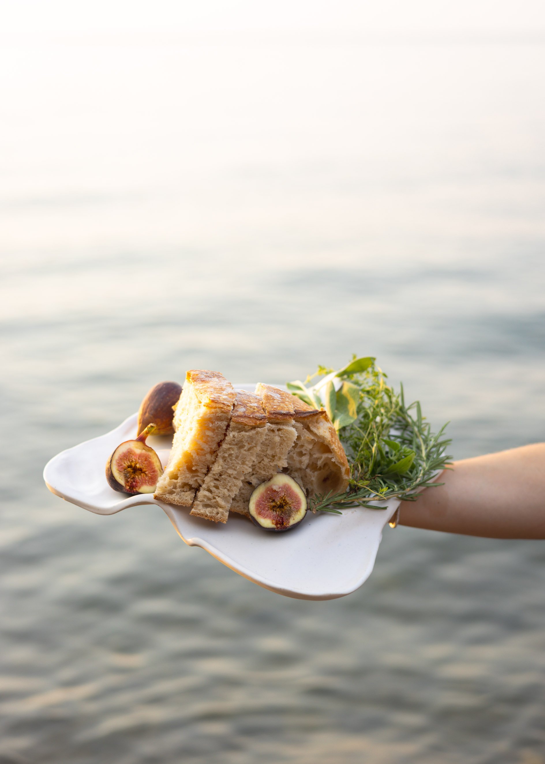 A slice of cake with figs and green herbs garnished on a white plate, held over a body of water with a cloudy sky in the background.