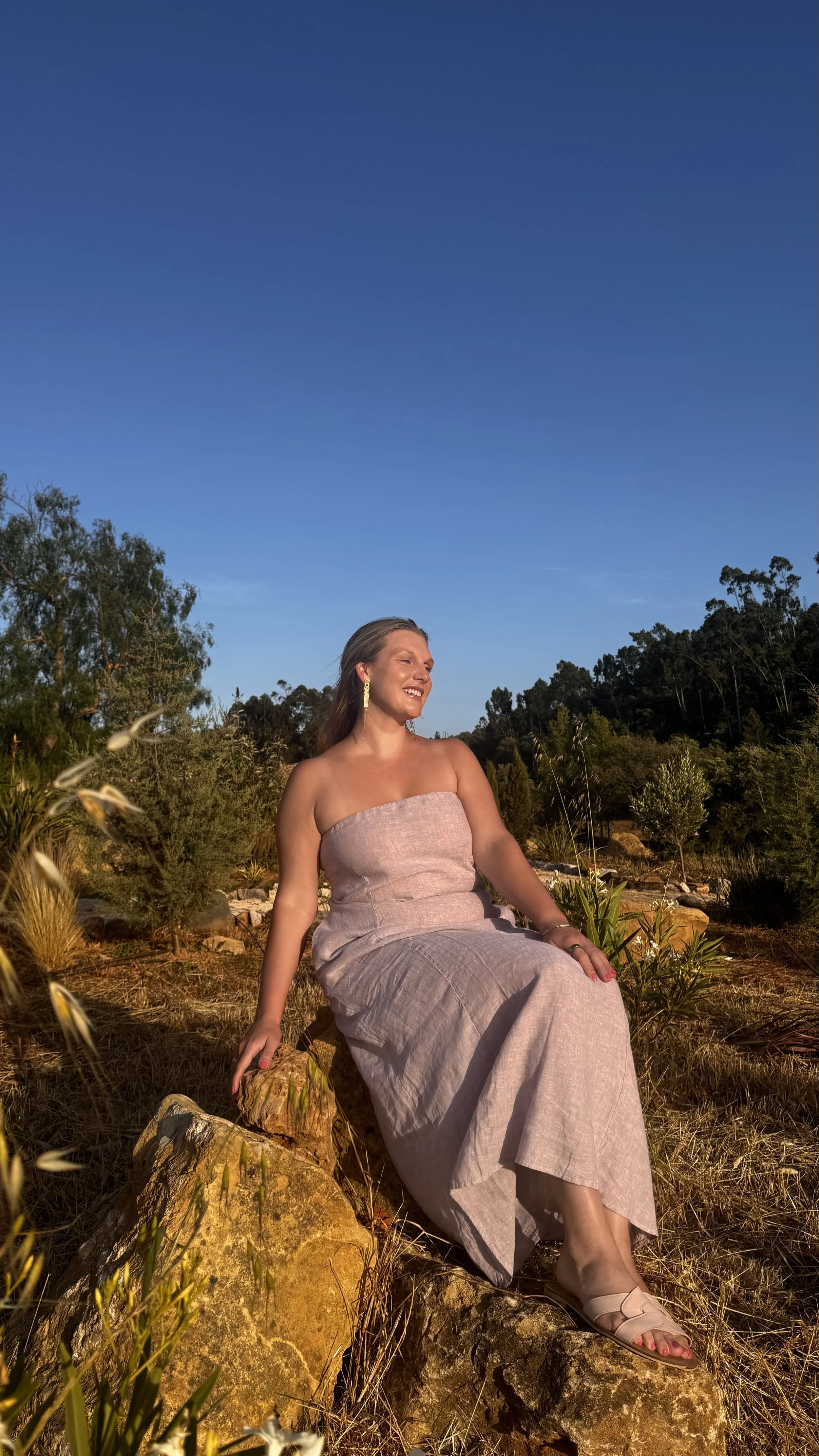 A woman sitting outdoors on a large rock, wearing a strapless dress and smiling, with a landscape background of trees and a clear blue sky.