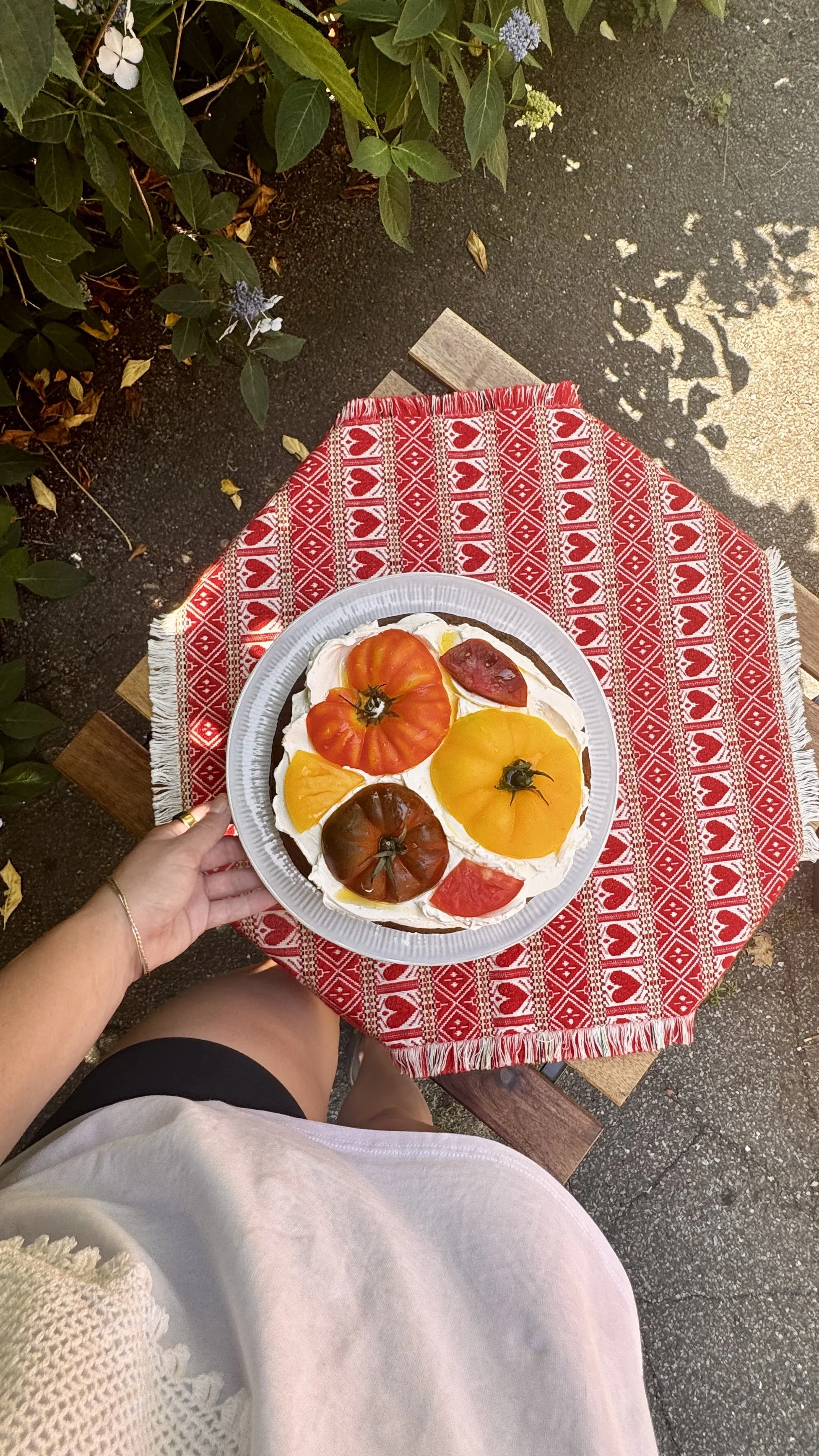 A person holding a fruit tart topped with heirloom tomatoes, sitting at a table with a red and white heart-patterned tablecloth outdoors.