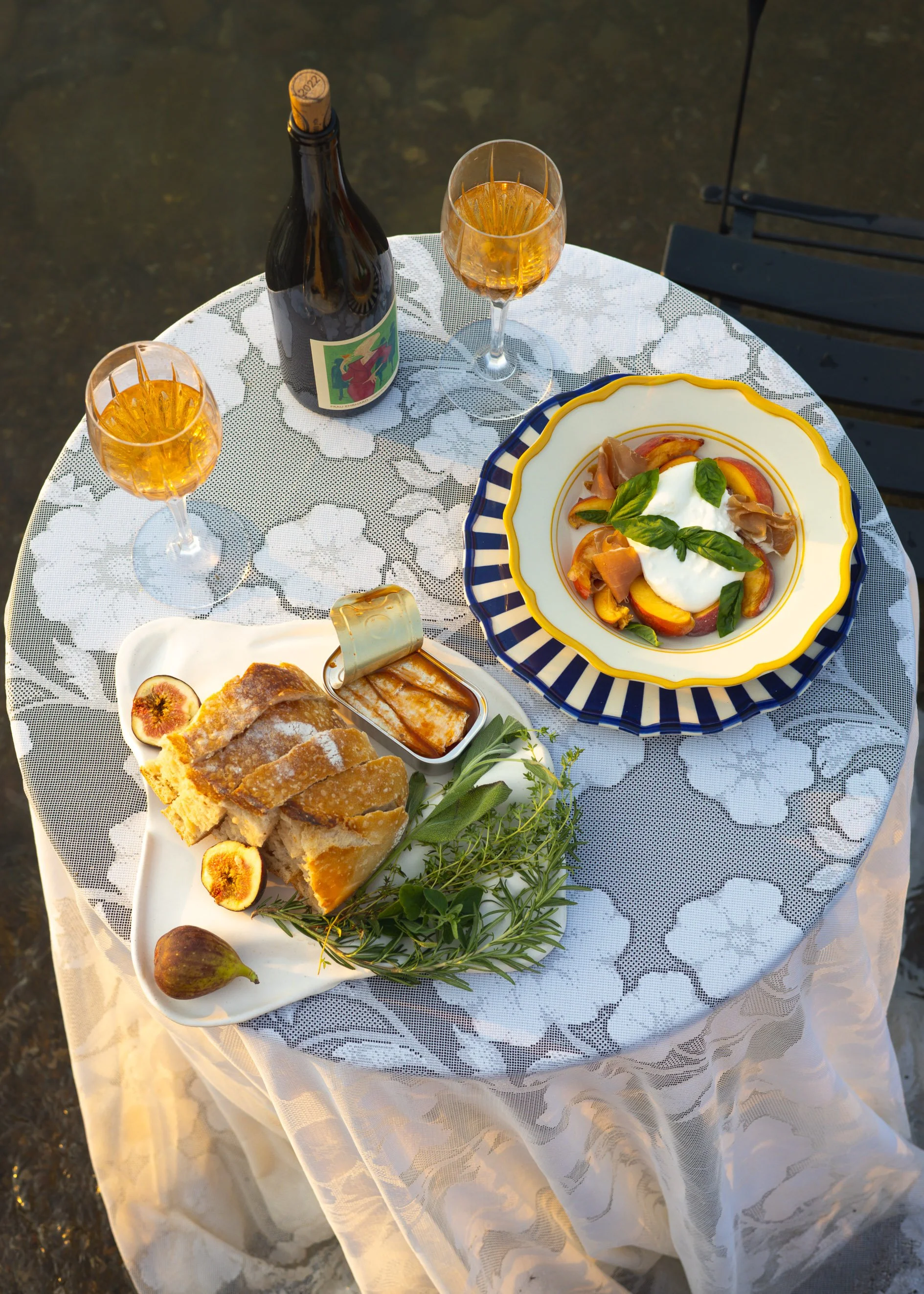 A round table with a white floral lace tablecloth set with a bottle of sparkling wine, two glasses of rosé, and a colorful salad with fresh herbs, cheese, and sliced fruit. There is also a plate with baked apple slices drizzled with caramel, figs, and sprigs of greenery.
