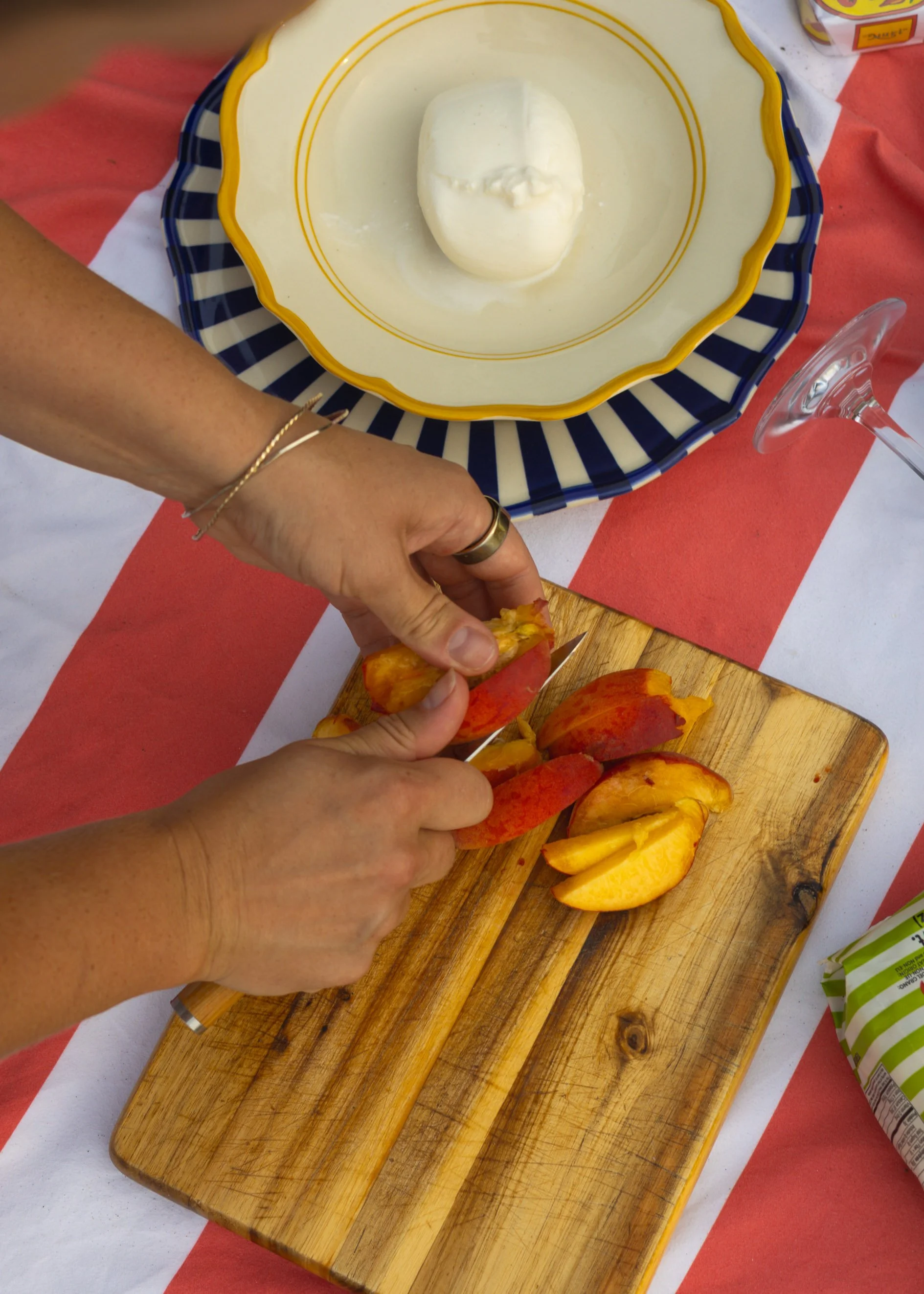 Person slicing a red and yellow peach on a wooden cutting board, with peeled peach slices nearby, on a striped serving platter with a bowl of mayonnaise in the background.