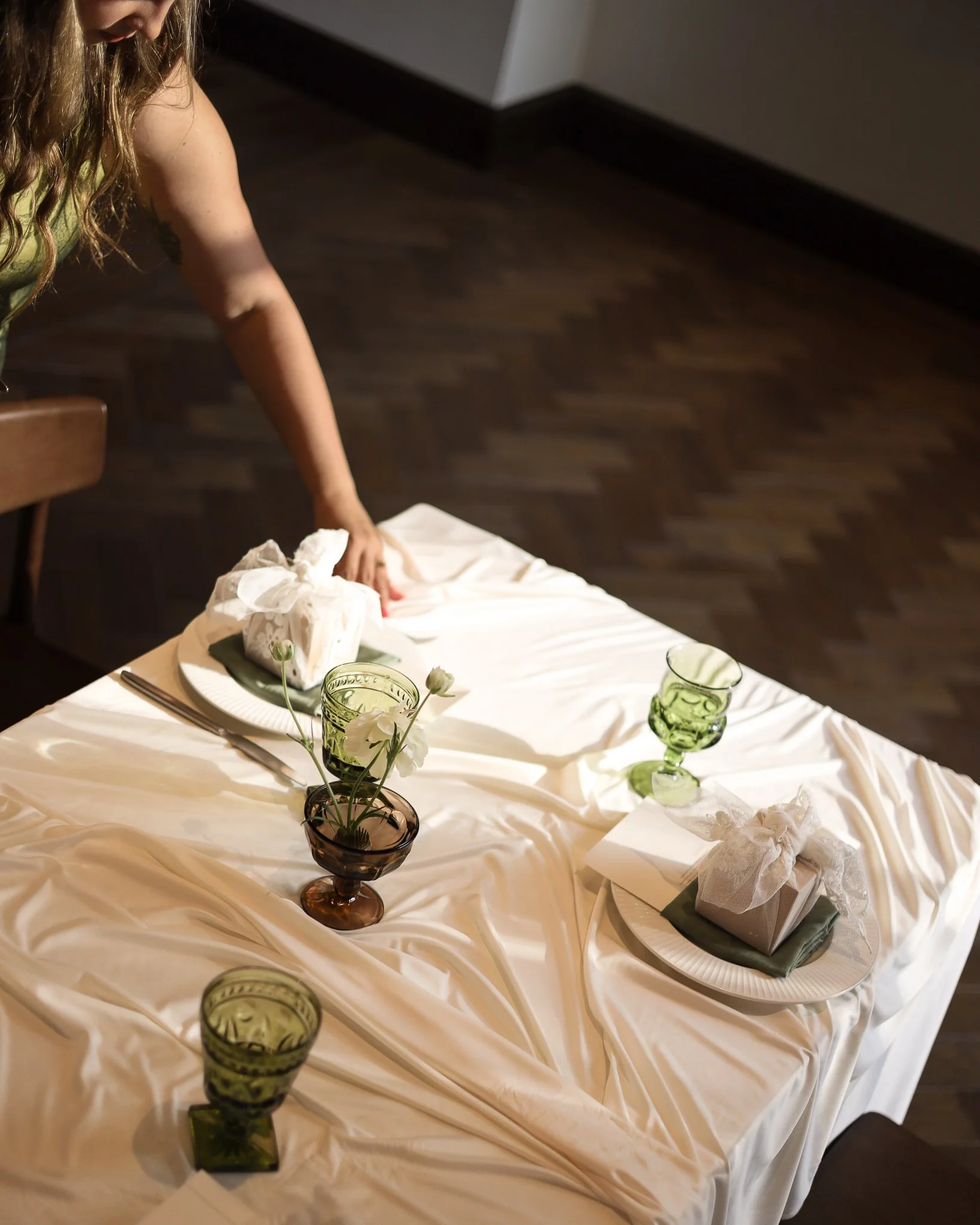 A table set for a celebration with a white tablecloth, green glasses, and a flower arrangement in a small dark vase. There are wrapped gifts on small plates, and the table is decorated with flowers and greenery.
