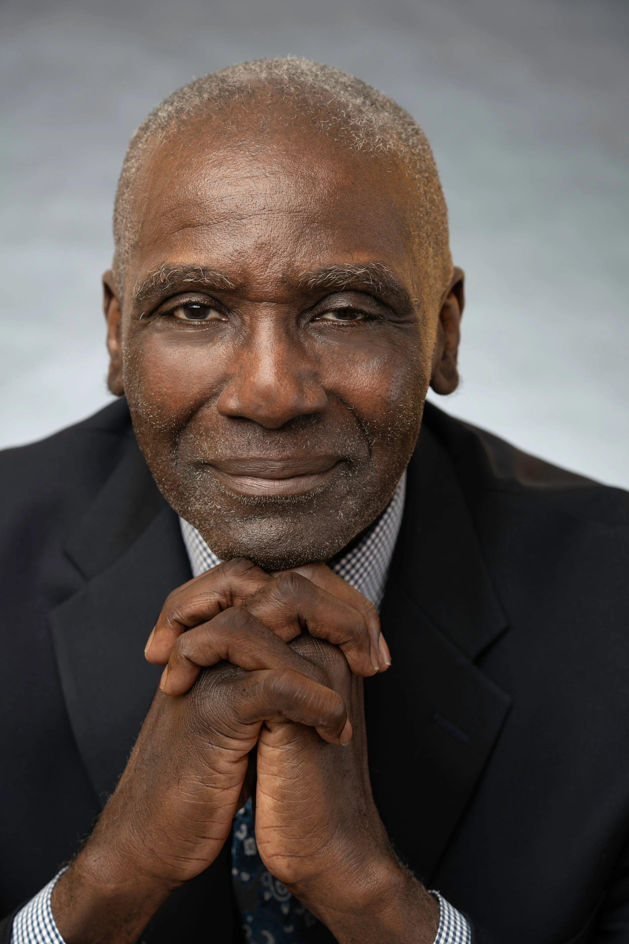 Close-up of an older Black man with a bald head and gray hair on the sides, wearing a dark suit and a checkered shirt, with his hands clasped under his chin, against a blurred gray background.