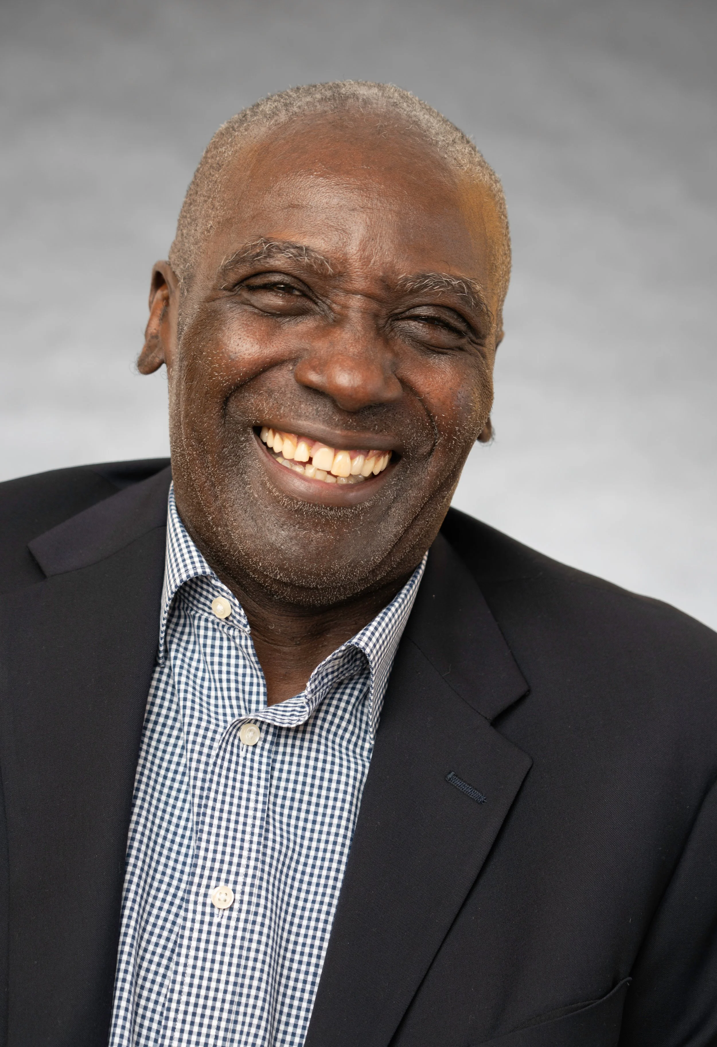 Close-up of an elderly African American man smiling, wearing a dark suit and a checkered shirt, against a plain gray background.