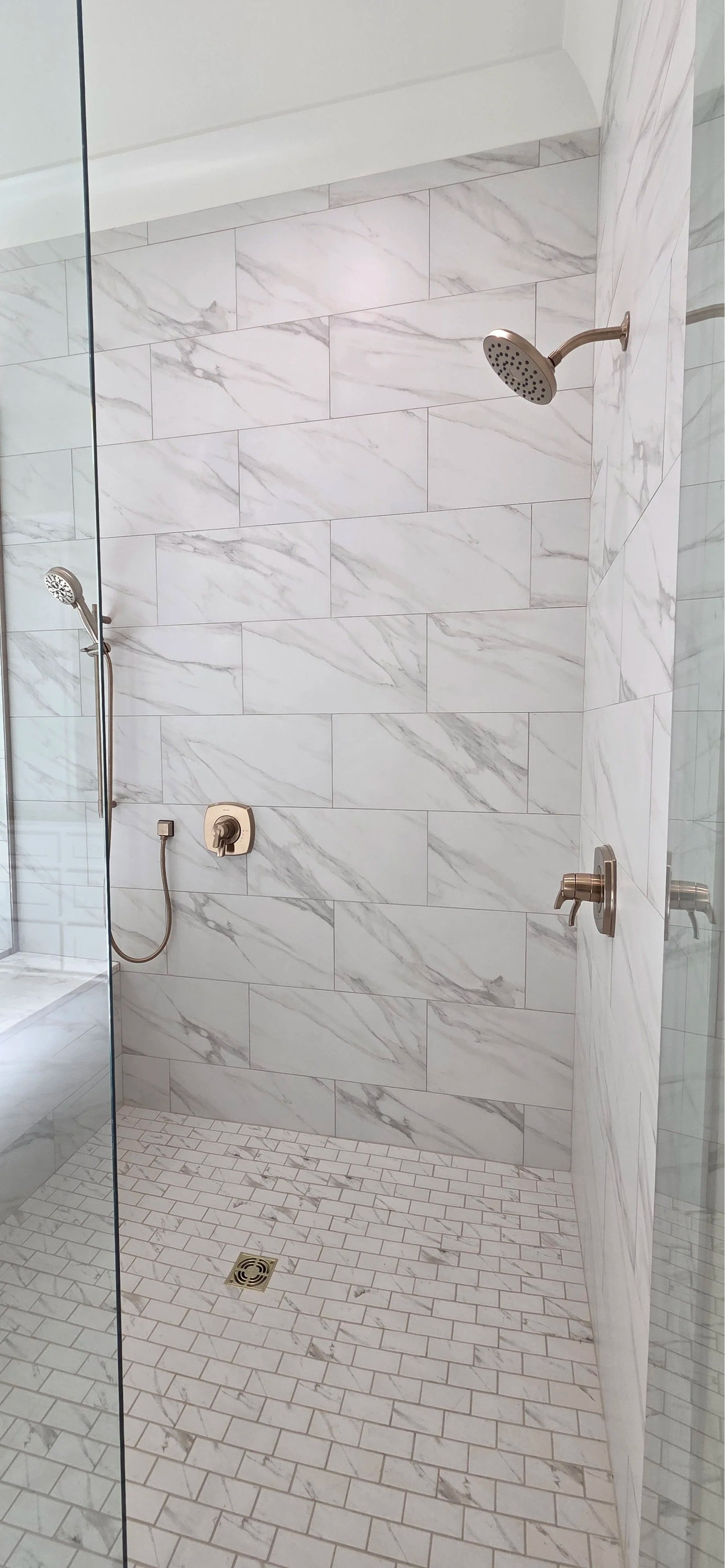 A walk-in shower with white marble tiles on the walls and small white tiles on the floor, featuring a rain showerhead and a hand-held showerhead.