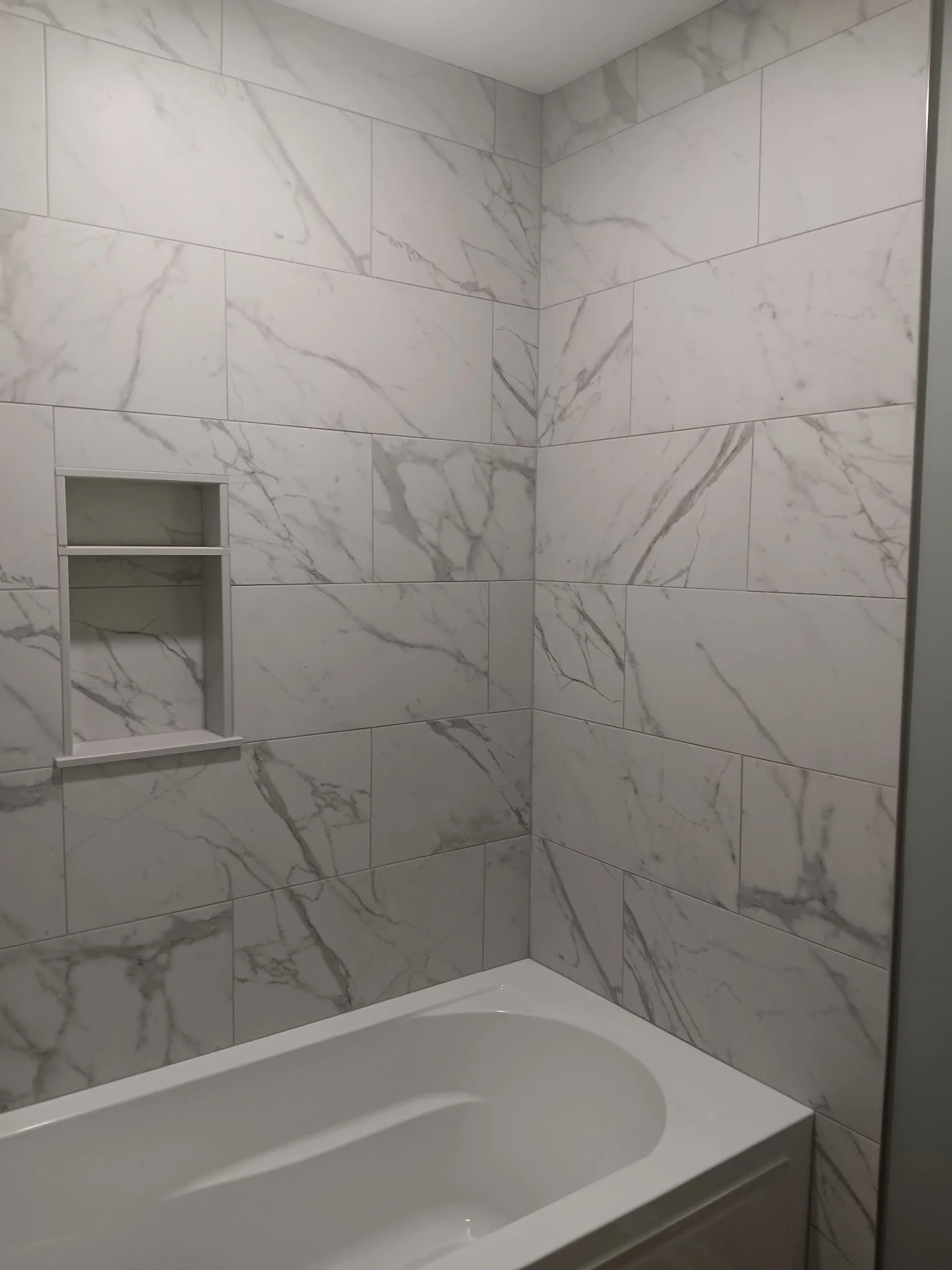 A close-up view of a bathroom corner with white marble tiles featuring gray veining, a built-in shower niche, and a partial view of a white bathtub.