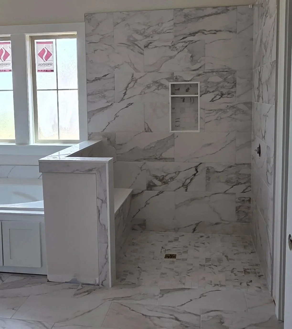 A marble tiled shower area with a small built-in bench, a recessed niche, and a window providing natural light, in a bathroom under construction.