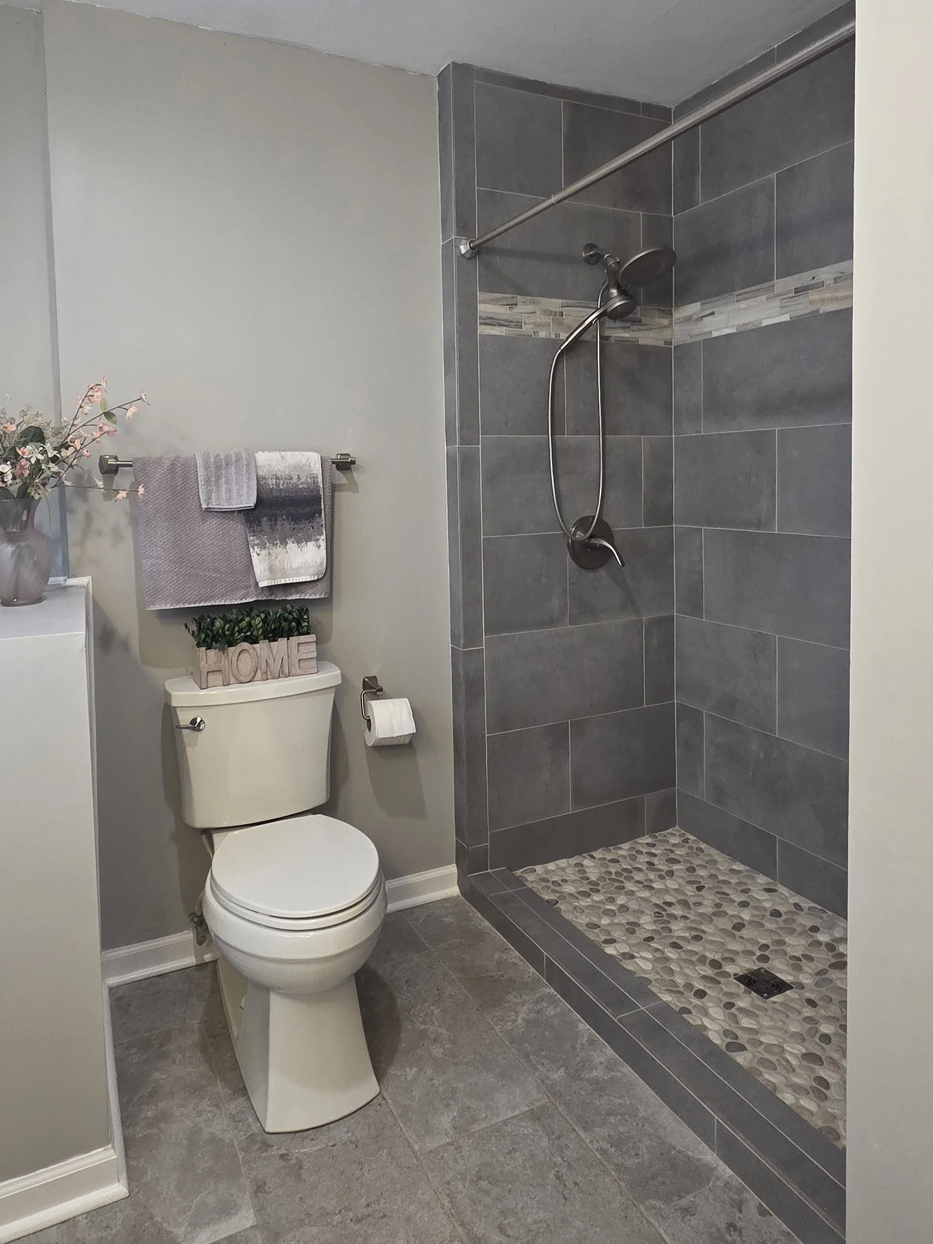 Bathroom with a toilet, a shower area with gray tiles, and decorative stones on the shower floor. A towel rail with towels, a small shelf with a plant and decorative 'HOME' sign.