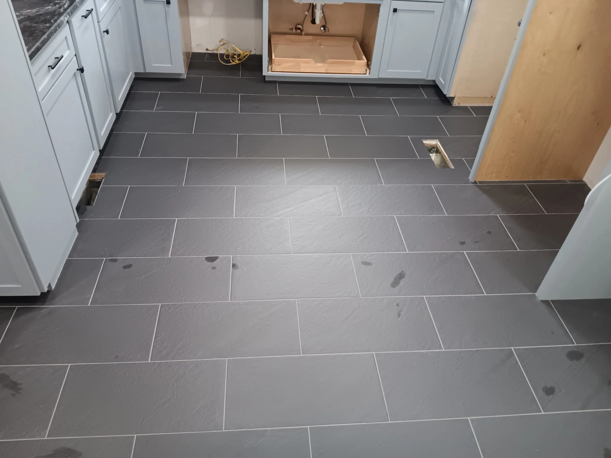 A kitchen under renovation showing gray tile flooring, white cabinets, and an open cabinet with plumbing pipes and a wooden box inside. There are two floor vents, with one near the center and the other behind a cabinet.