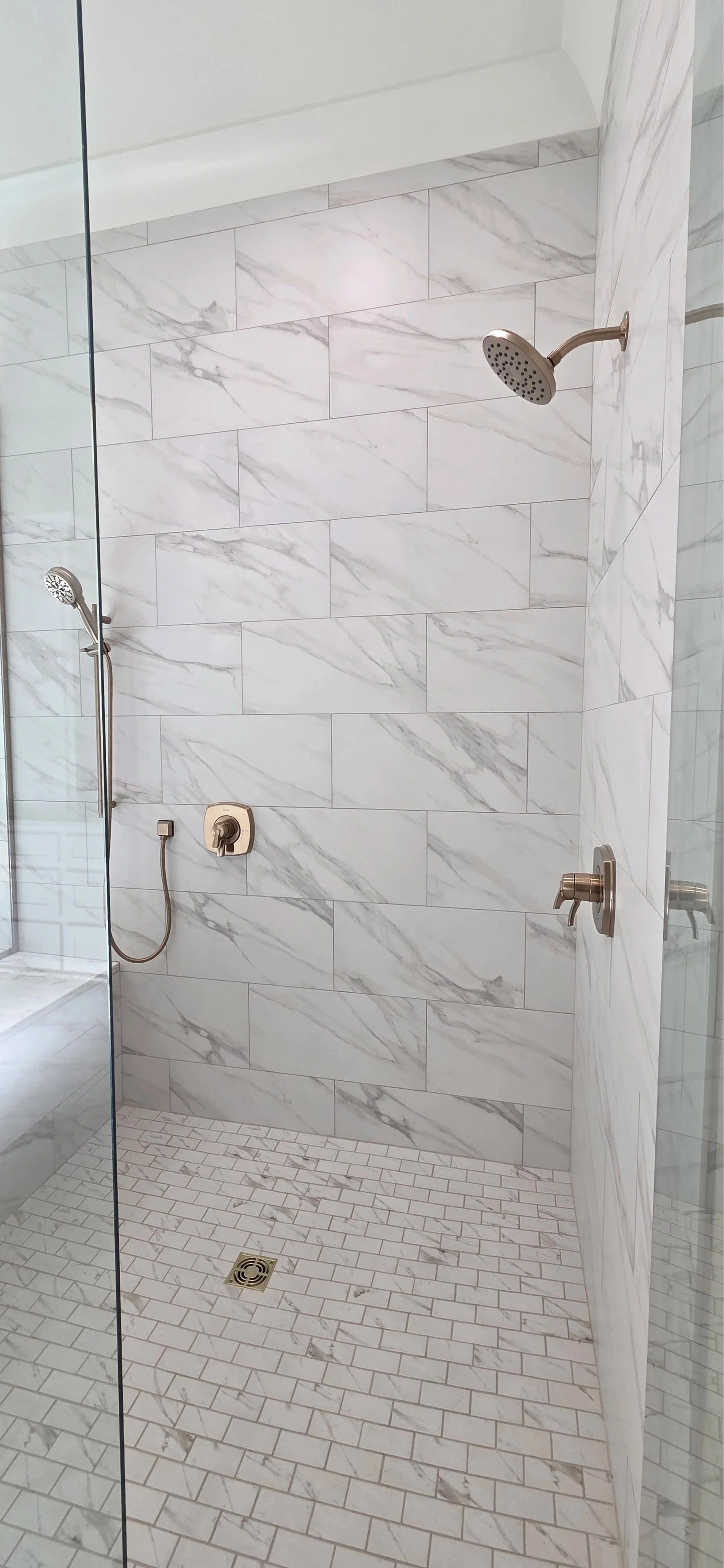 A modern walk-in shower with white marble tiles, a rainfall shower head, a handheld shower, and a bronze wall-mounted faucet.