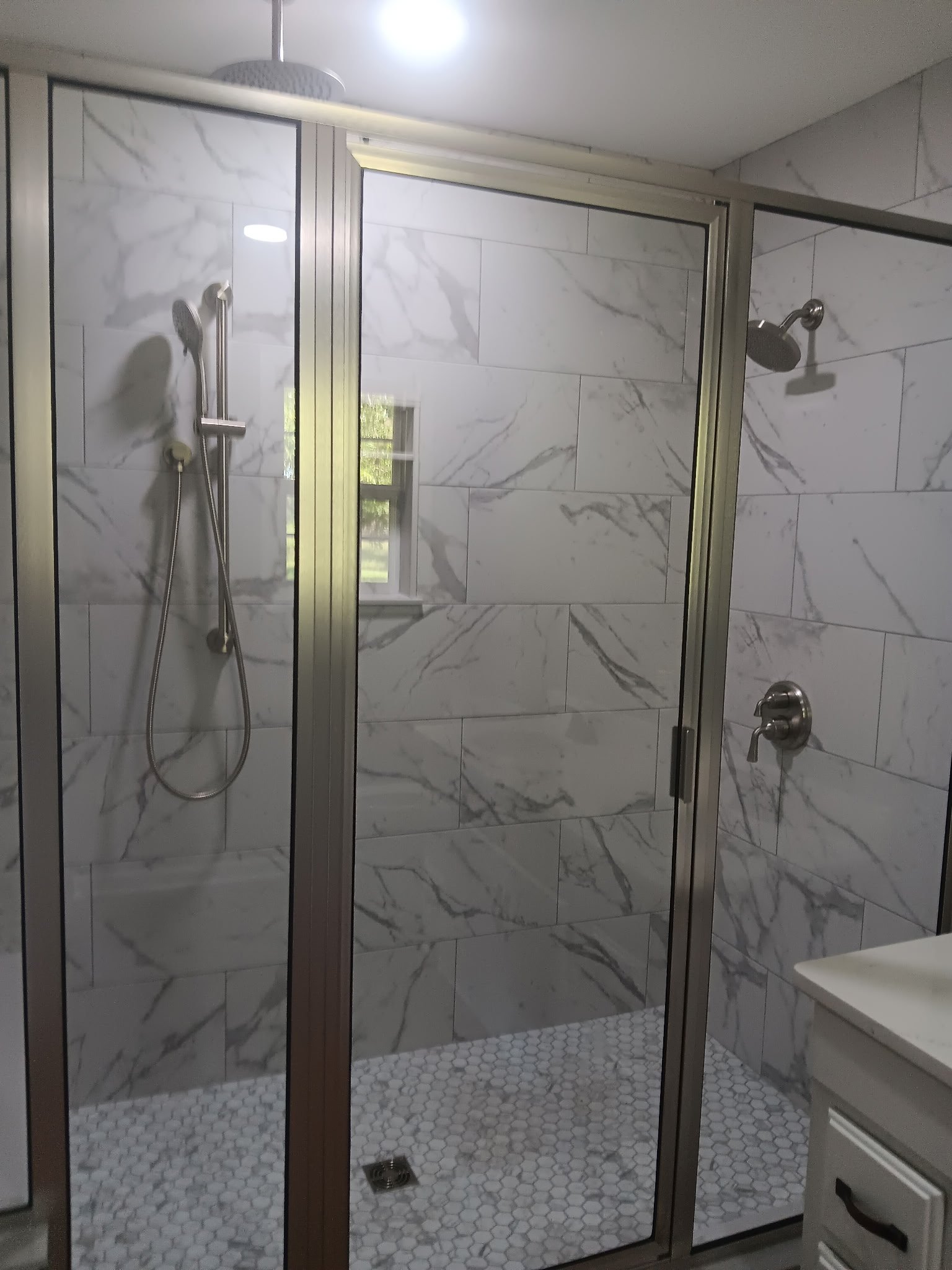 A walk-in shower with white marble tiles on the walls and small white hexagon tiles on the floor. The shower has two showerheads, one on each side, with a glass door and metal framing. A window is visible through the glass door, letting in natural light.
