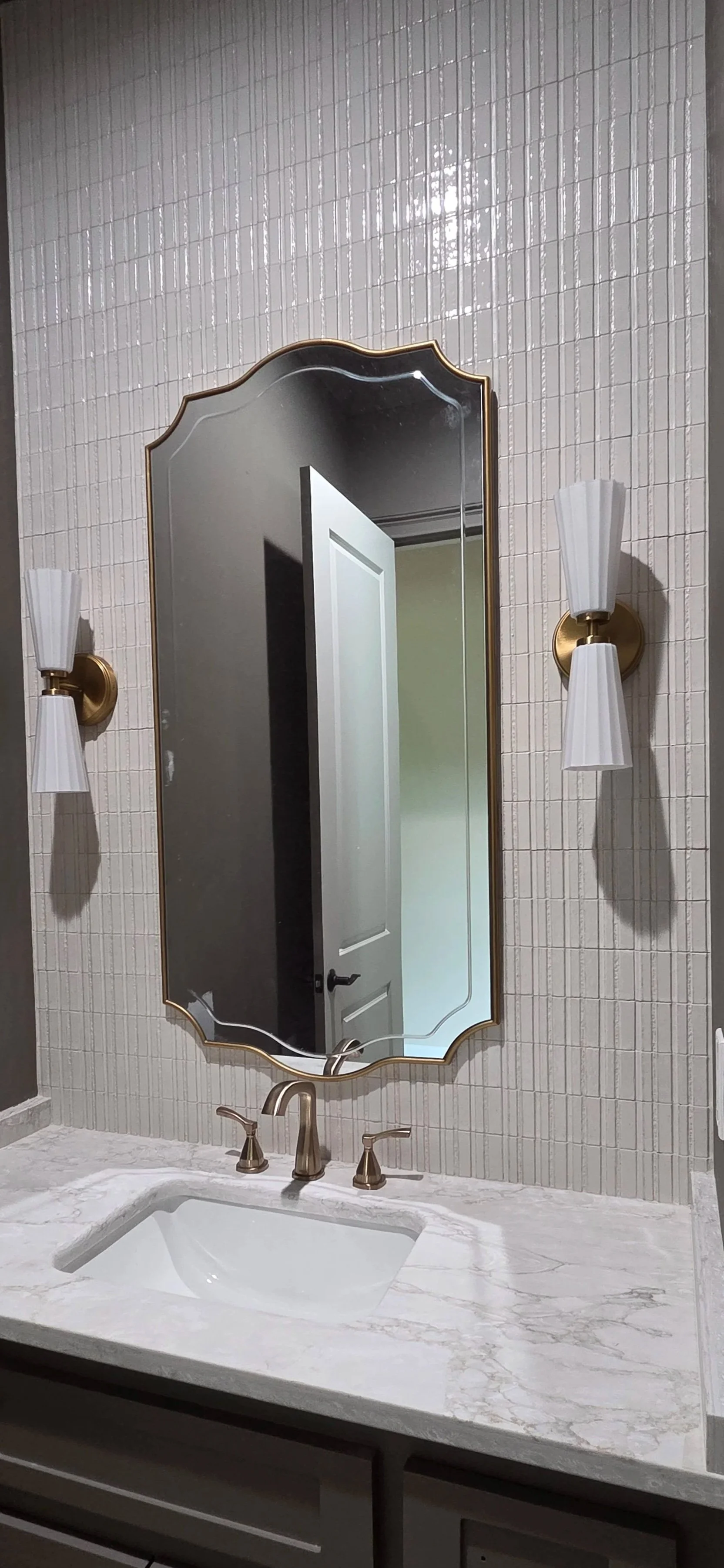 Bathroom with a marble countertop sink, brushed gold faucet, an oval mirror with gold trim, and two wall-mounted light fixtures with white shades