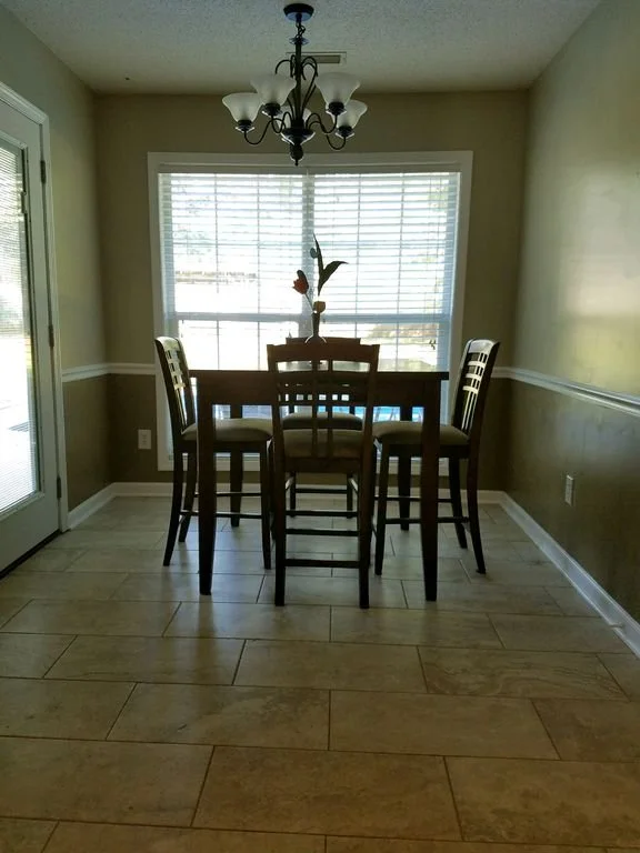 A dining room with a wooden table and six chairs, a window with blinds, a chandelier hanging from the ceiling, and beige walls with white trim.