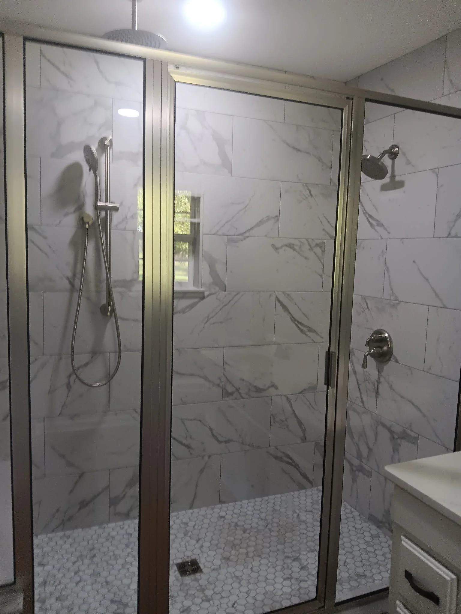 A modern walk-in shower with a clear glass enclosure, marble tile walls, two shower heads, and white hexagonal floor tiles.