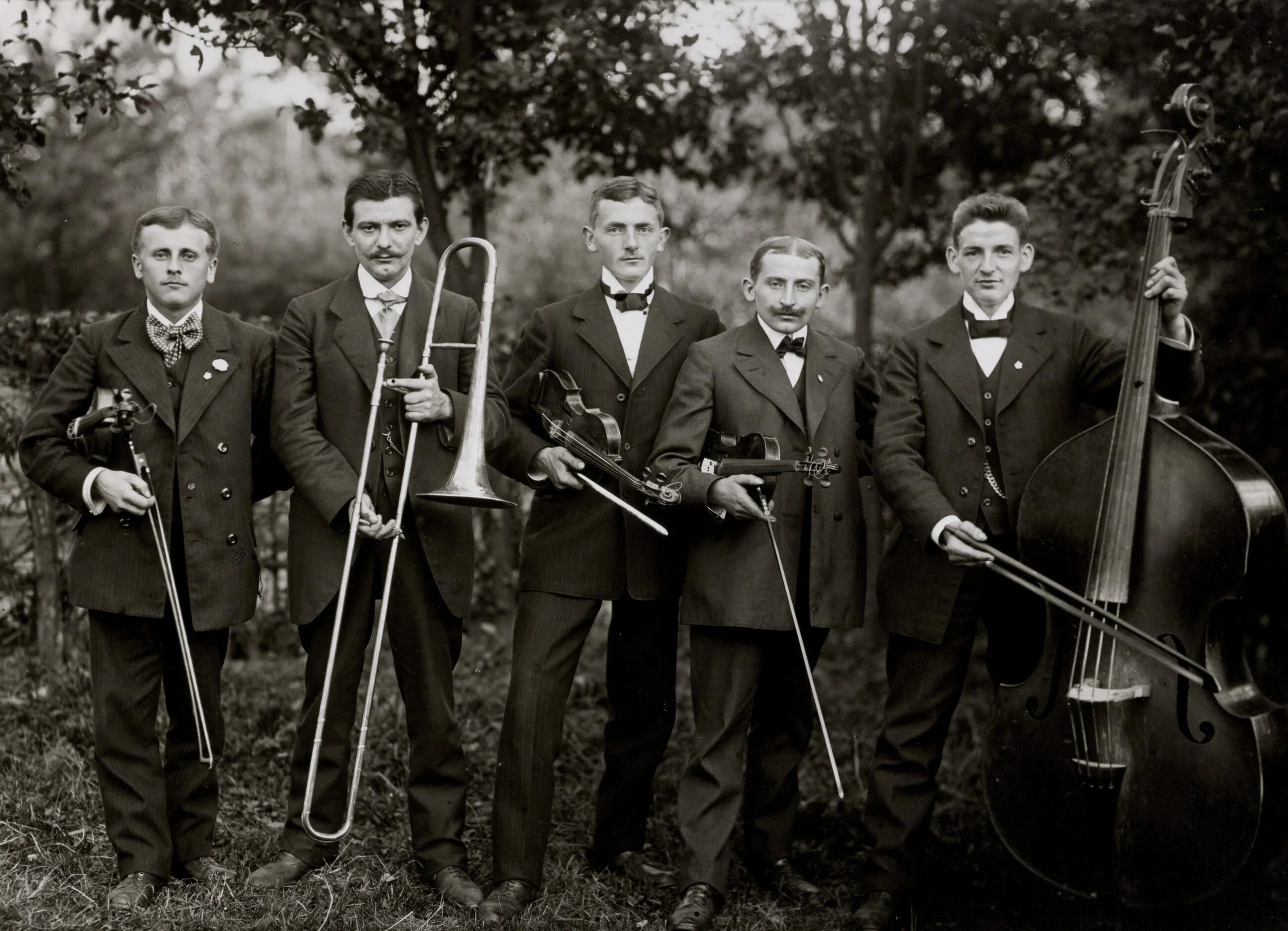 A black and white photograph of five men dressed in formal suits with bow ties, holding musical instruments, standing outdoors in front of trees.