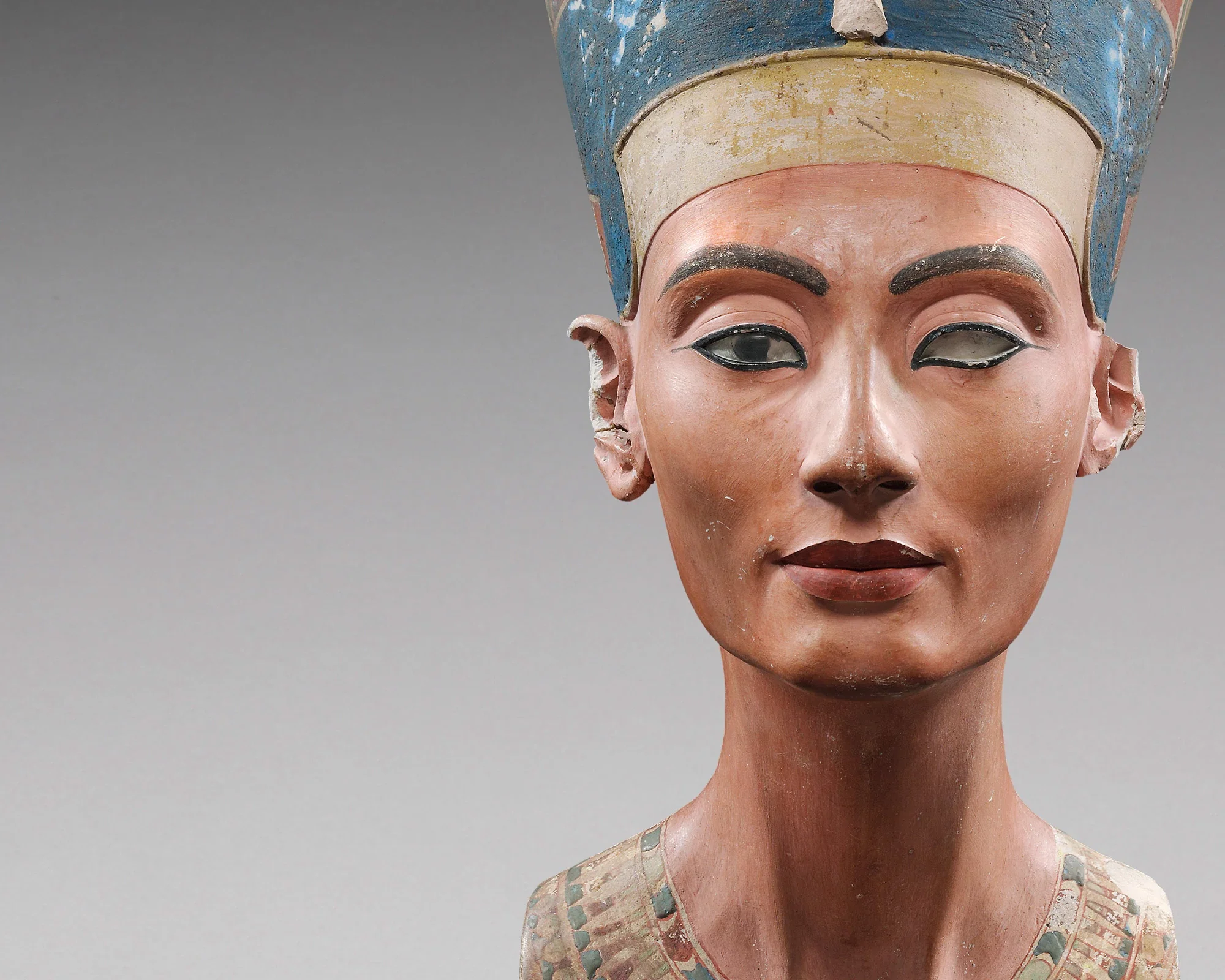 Close-up of an ancient Egyptian painted wooden bust of a woman with black eyebrows and lips, wearing a blue headdress with chipped paint, against a gray background.