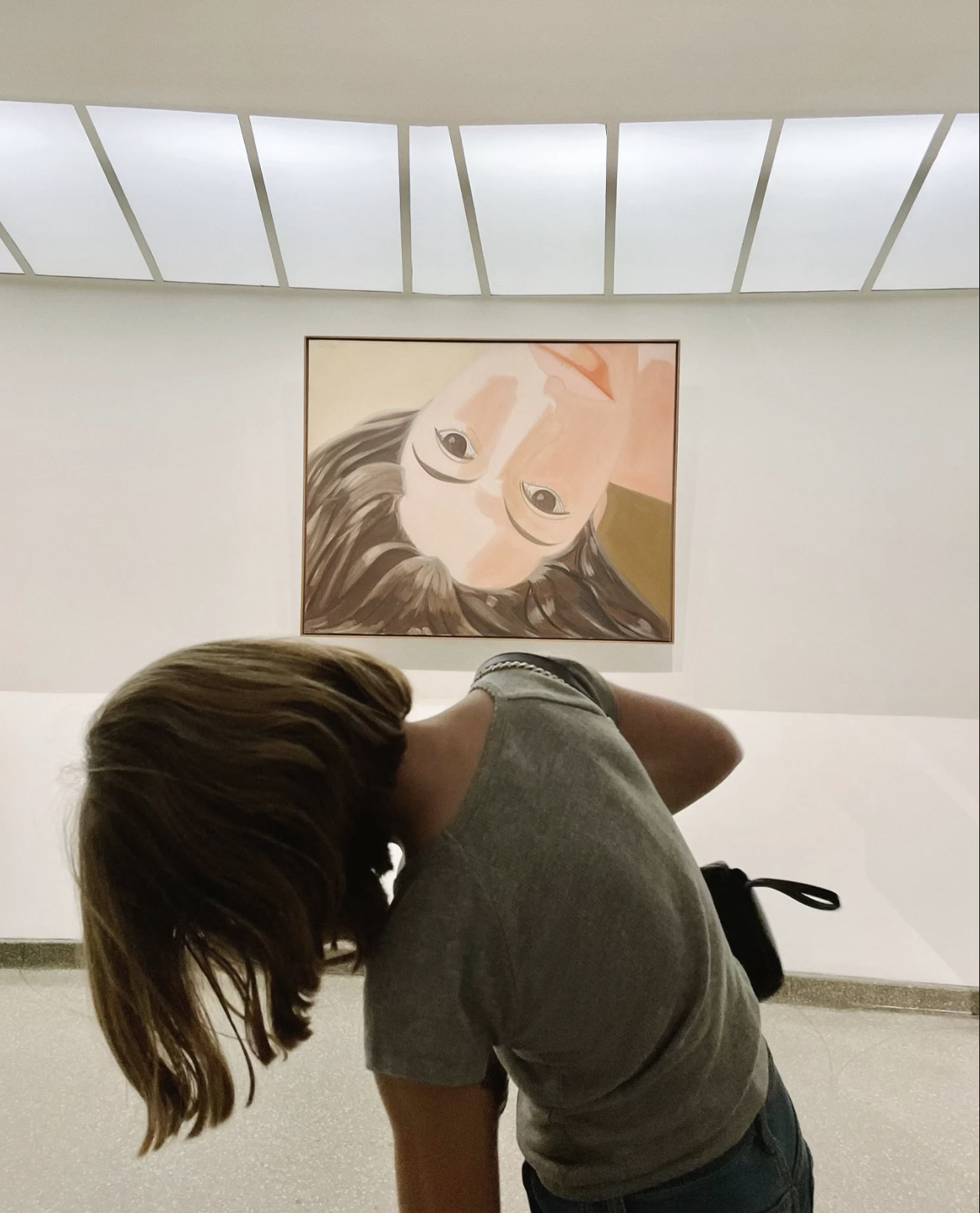 A person with brown hair is bent over, facing downward in front of a large wall portrait of a woman with glasses and long hair, inside an art gallery or museum.