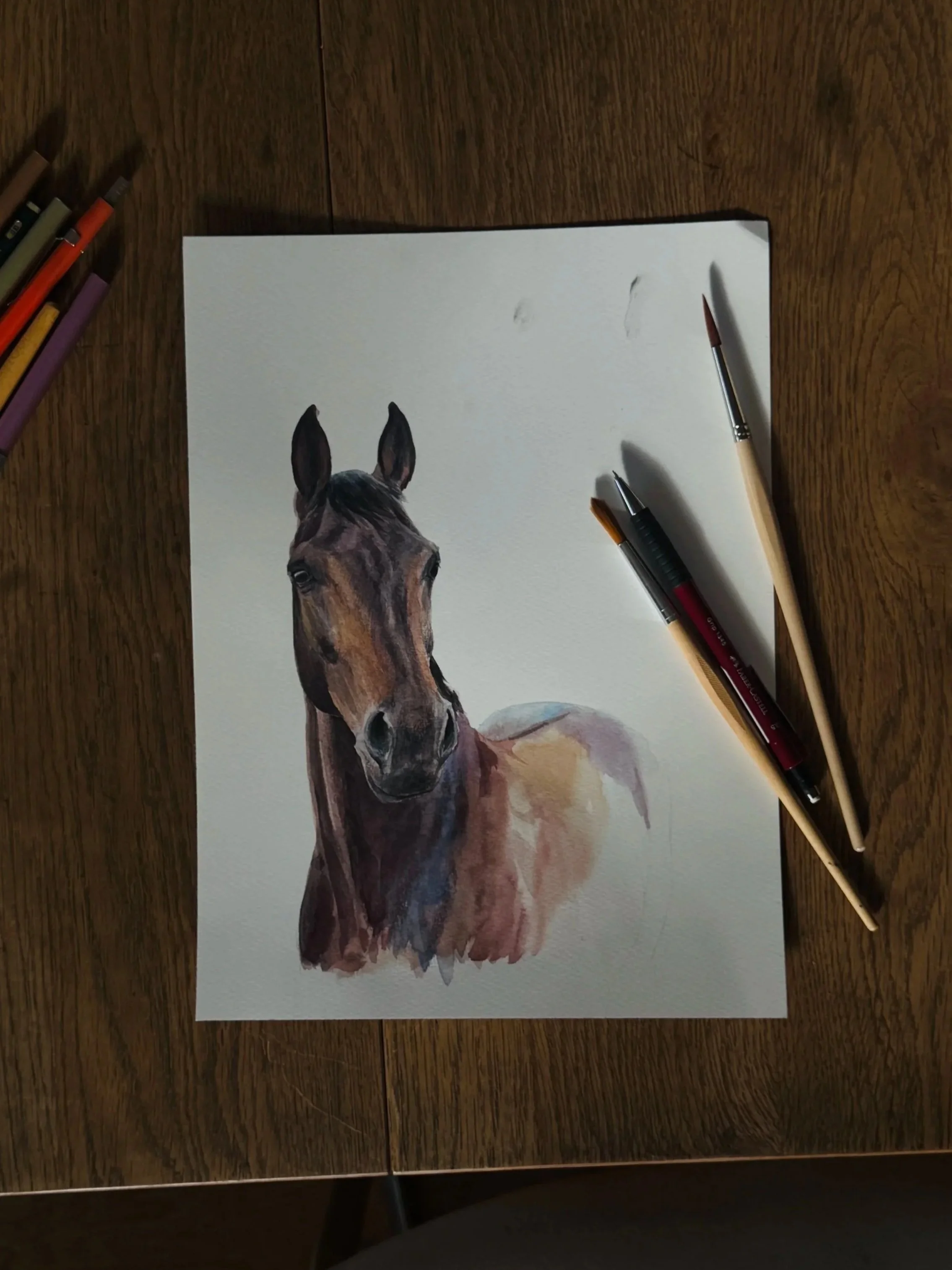 Watercolor painting of a horse's head and shoulders on white paper, with watercolor markers and paintbrushes beside it on a wooden table.
