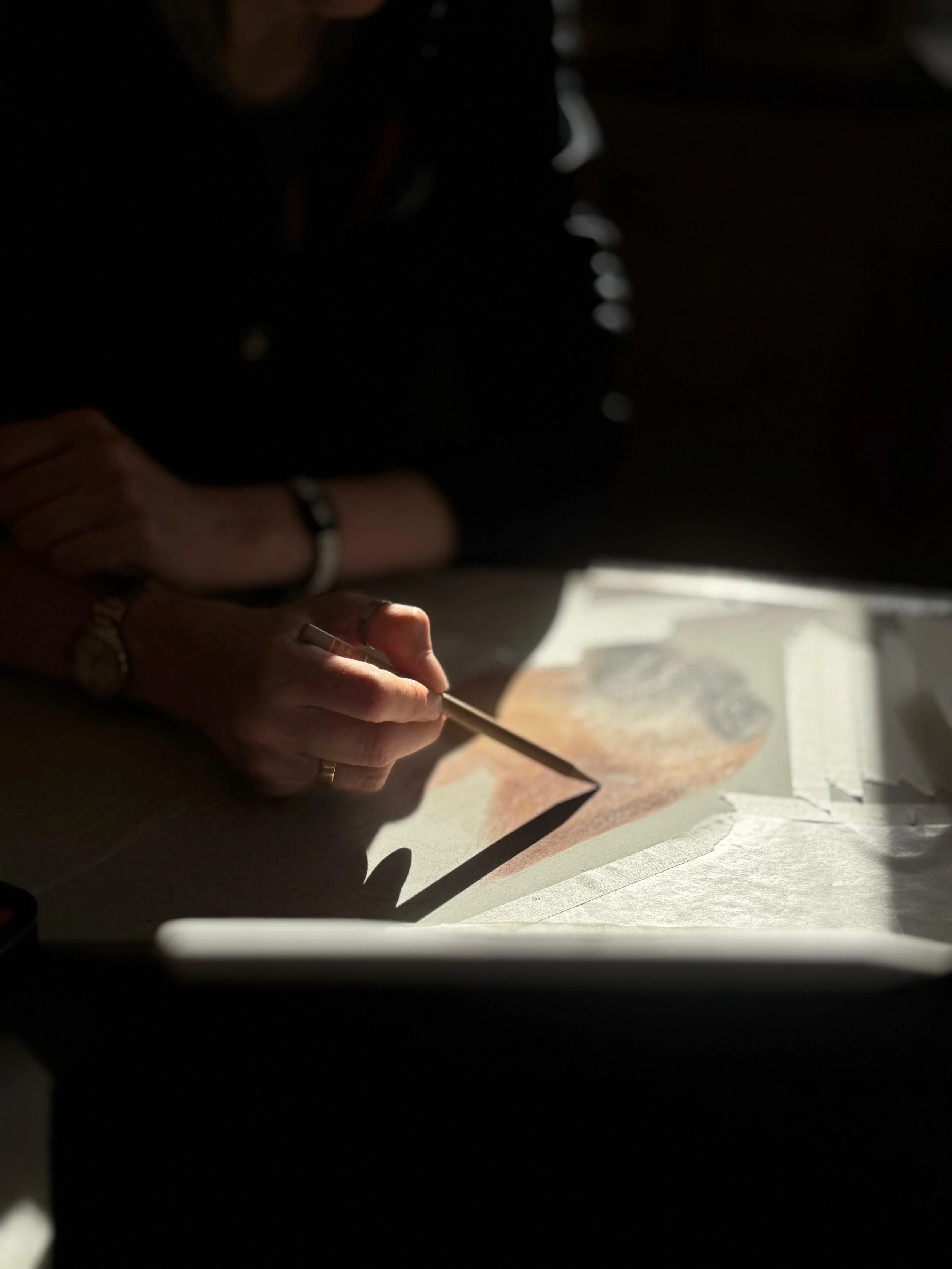 A person is working on a drawing or painting, using a brush or pencil, with sunlight illuminating their hands and the artwork on paper.
