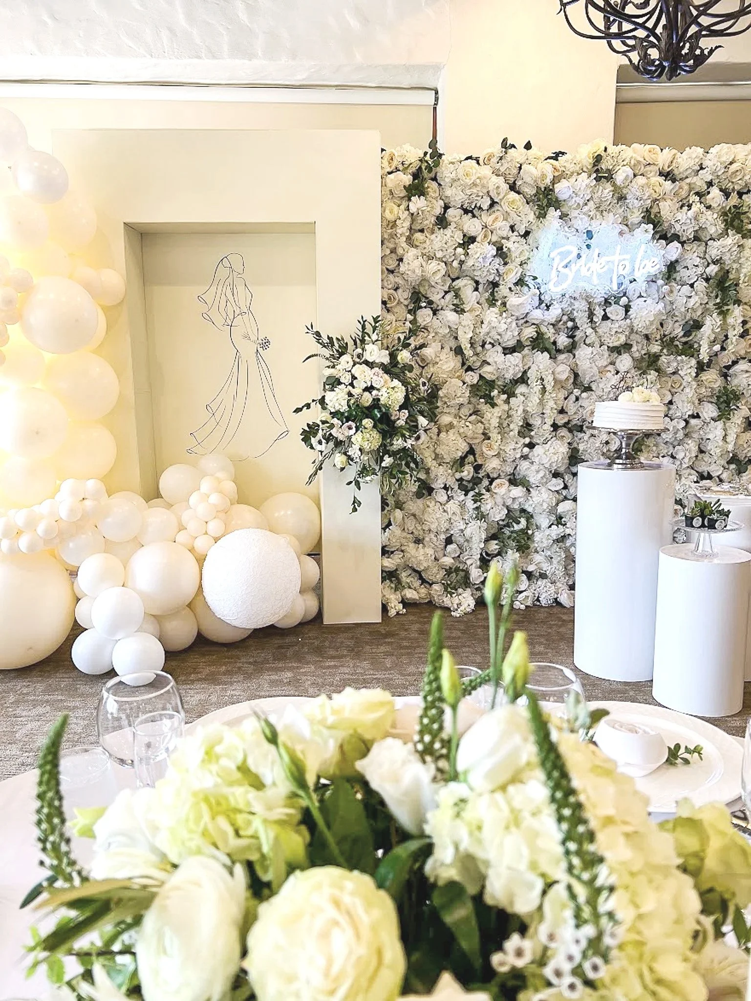 Indoor wedding reception area decorated with white flowers, balloons, and a neon sign that says 'Bride to Be'. There is a floral backdrop with a silhouette of a bride and a small table with a cake on it.