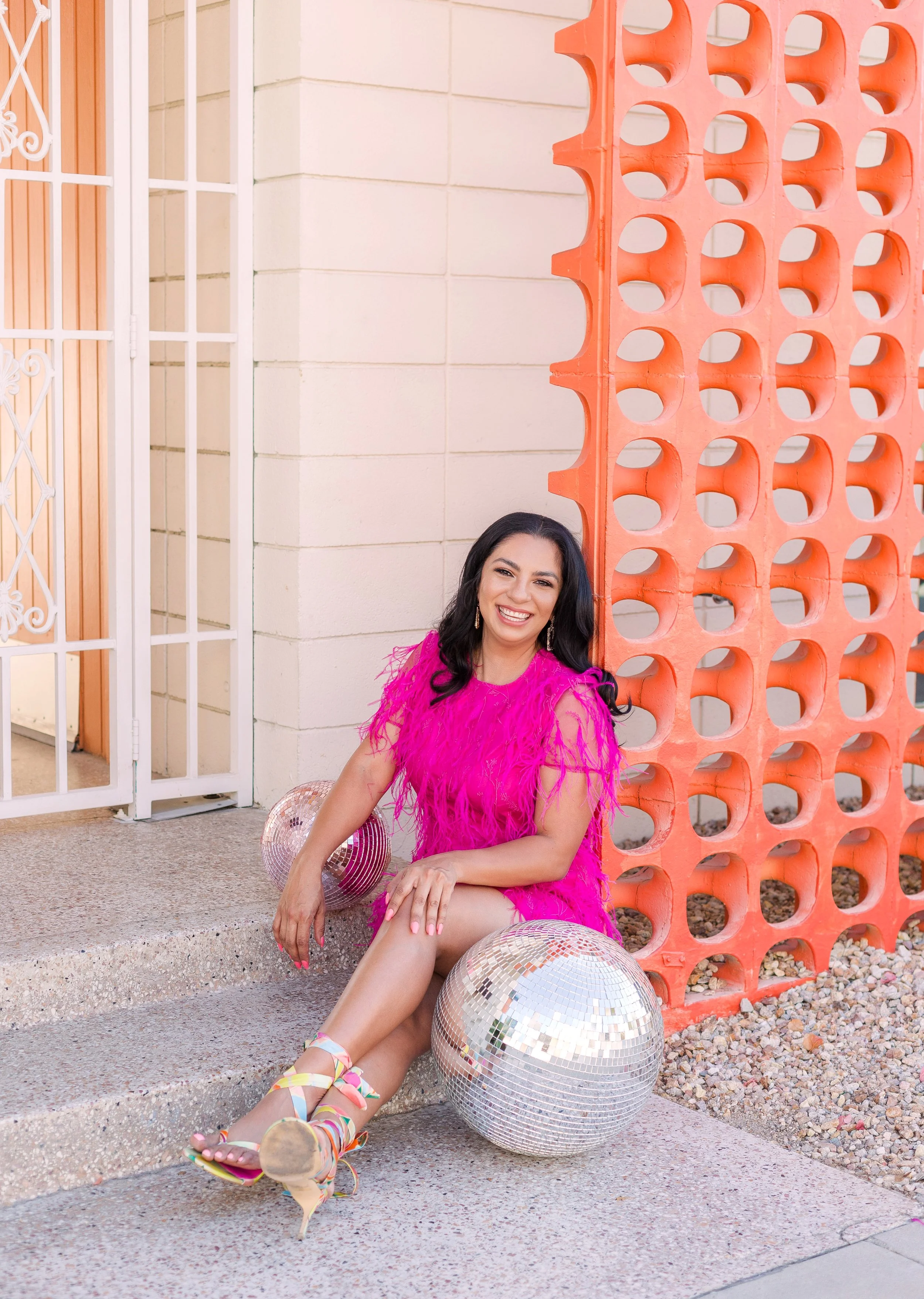 Woman in a bright pink feathered dress sitting on steps next to a silver disco ball and another mirrored ball, smiling at the camera, with an orange decorative siding and white brick wall in the background.