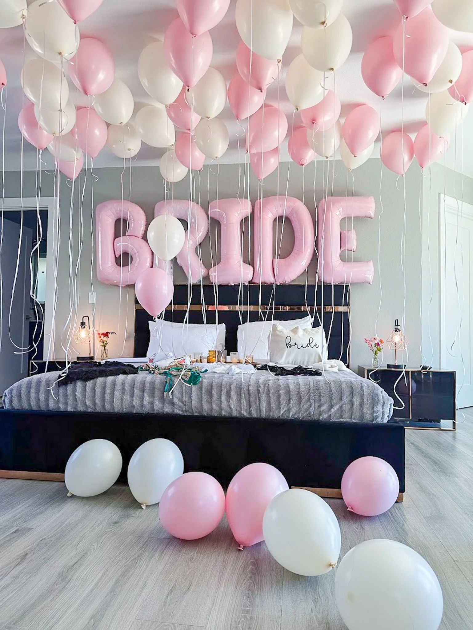 Decorated bedroom with pink and white balloons hanging from the ceiling, large pink 'LOVE' balloons on the wall, and additional pink and white balloons scattered on the bed and floor, indicating a romantic or celebratory occasion.