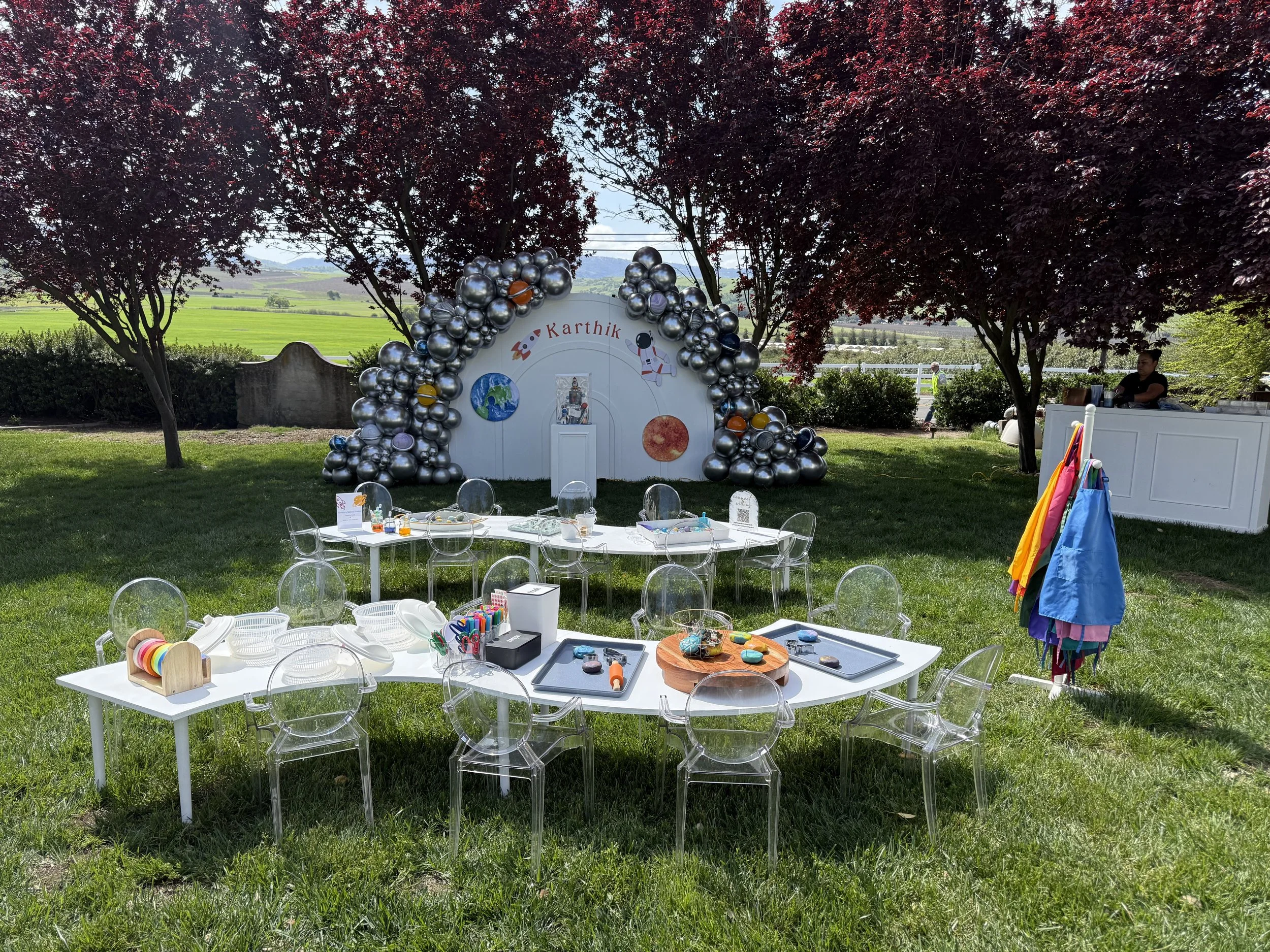 Outdoor space with white tables, clear chairs, and craft supplies, shaded by trees, with a decorated backdrop labeled 'Karthik' featuring balloons, planets, and space-themed decorations.