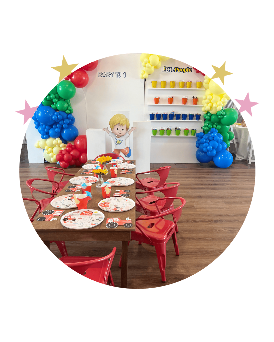 Decorated party area with red chairs around a wooden table, colorful paper plates and cups, balloon arch in red, yellow, green, blue, and purple, cartoon child illustration on the wall, and shelves with colorful cups labeled 'Little People' for a children's birthday celebration.