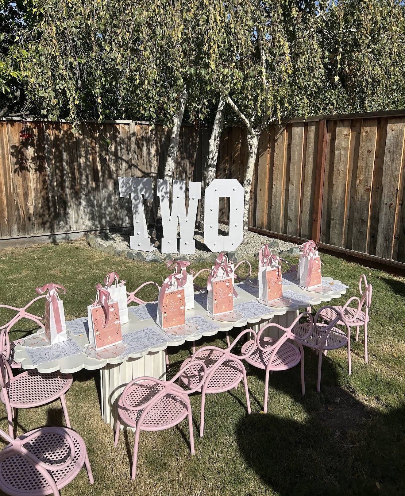 Decorated outdoor area for a celebration with a long white table surrounded by pink chairs. The table has pink gift bags with ribbons. Behind the table, there is a wooden fence with large white letters spelling 'TWO' and a tree with green leaves.
