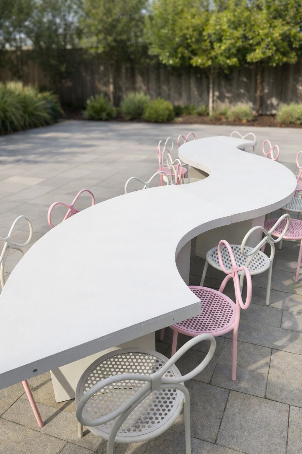 Outdoor patio with curved white table surrounded by pastel pink and white chairs, with trees and bushes in the background.